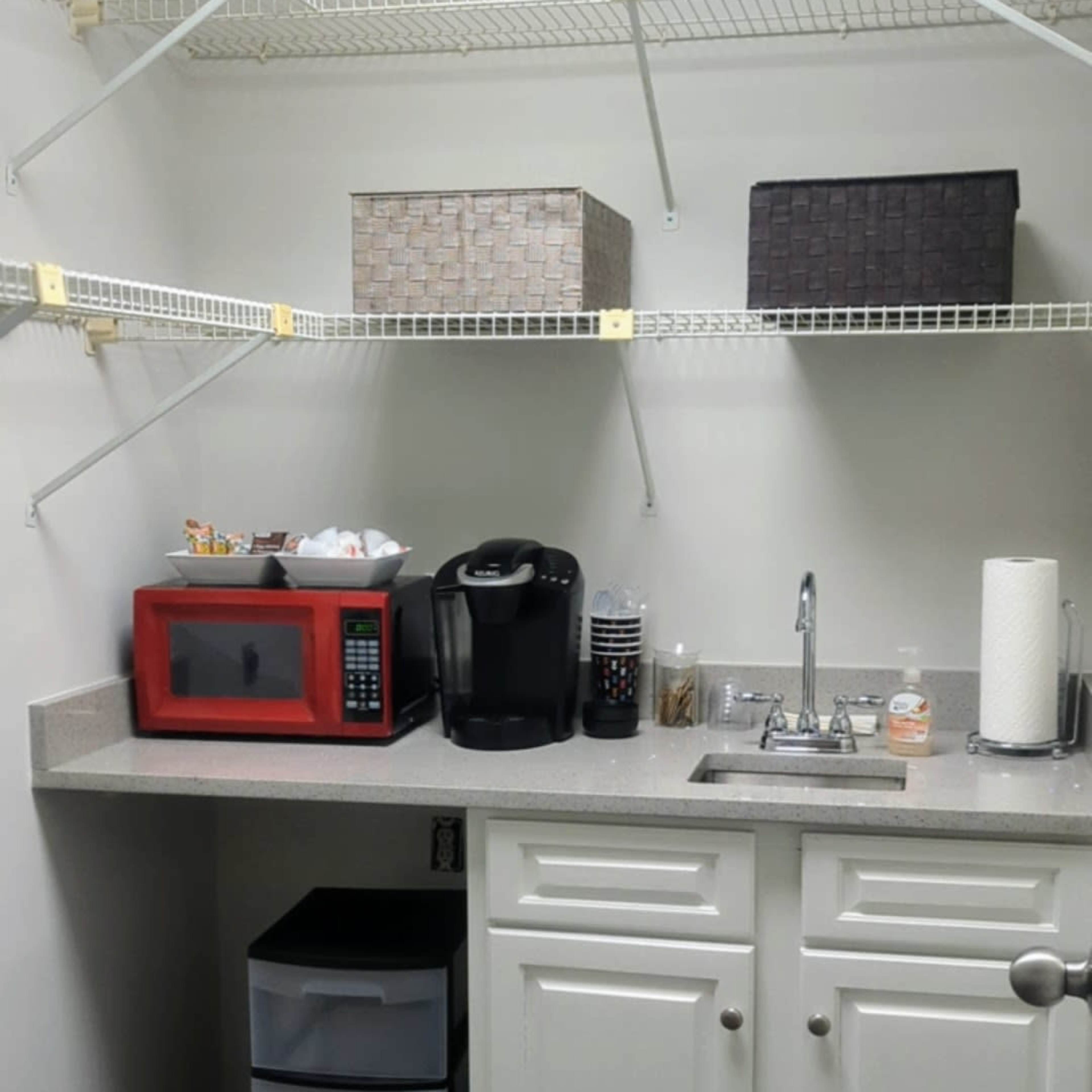A small storage room with shelves, a countertop that holds a microwave, a coffee maker, and a sink, along with a set of stacked drawers and various storage boxes.