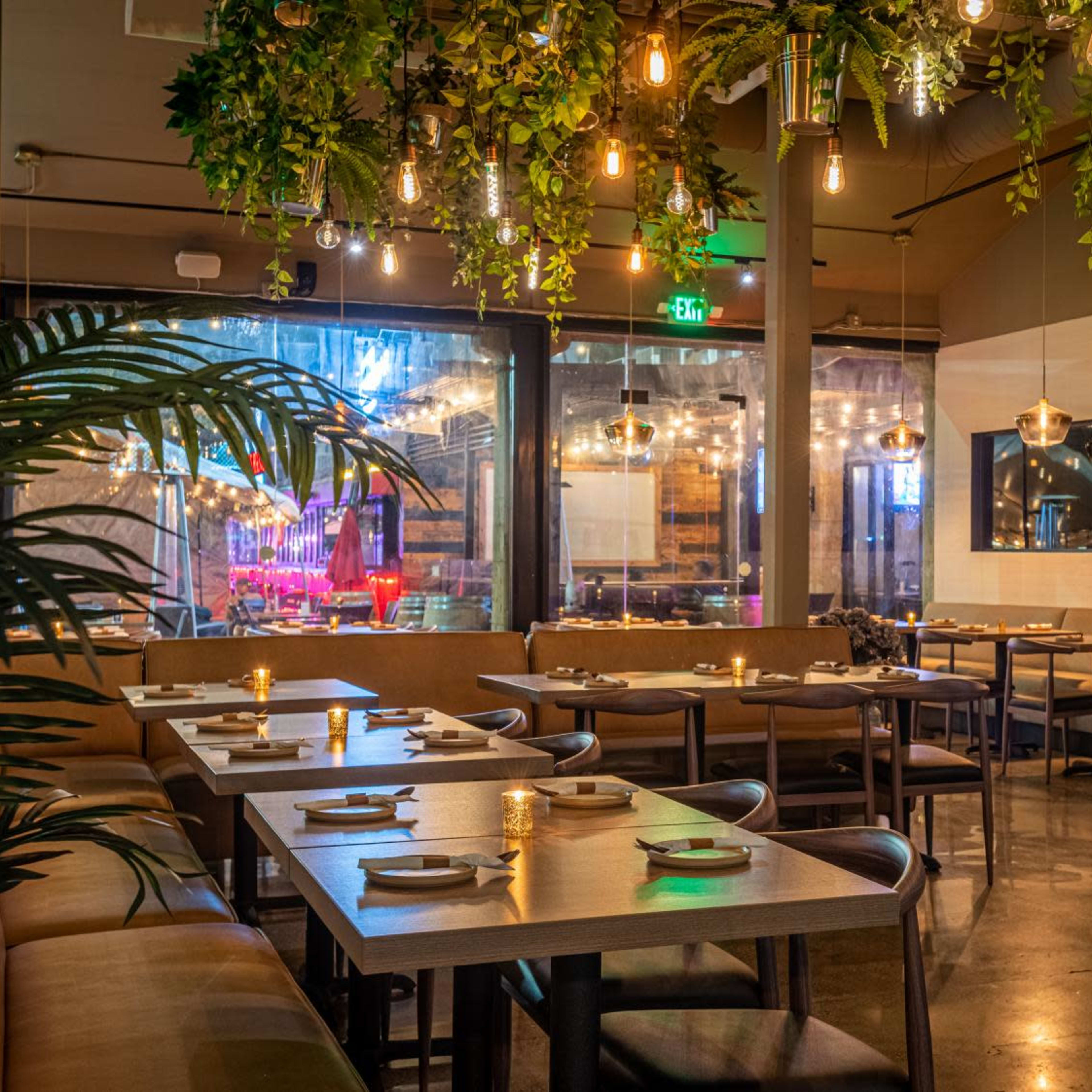 A modern restaurant interior with wooden tables set for dining, surrounded by decorative plants and ambient lighting.