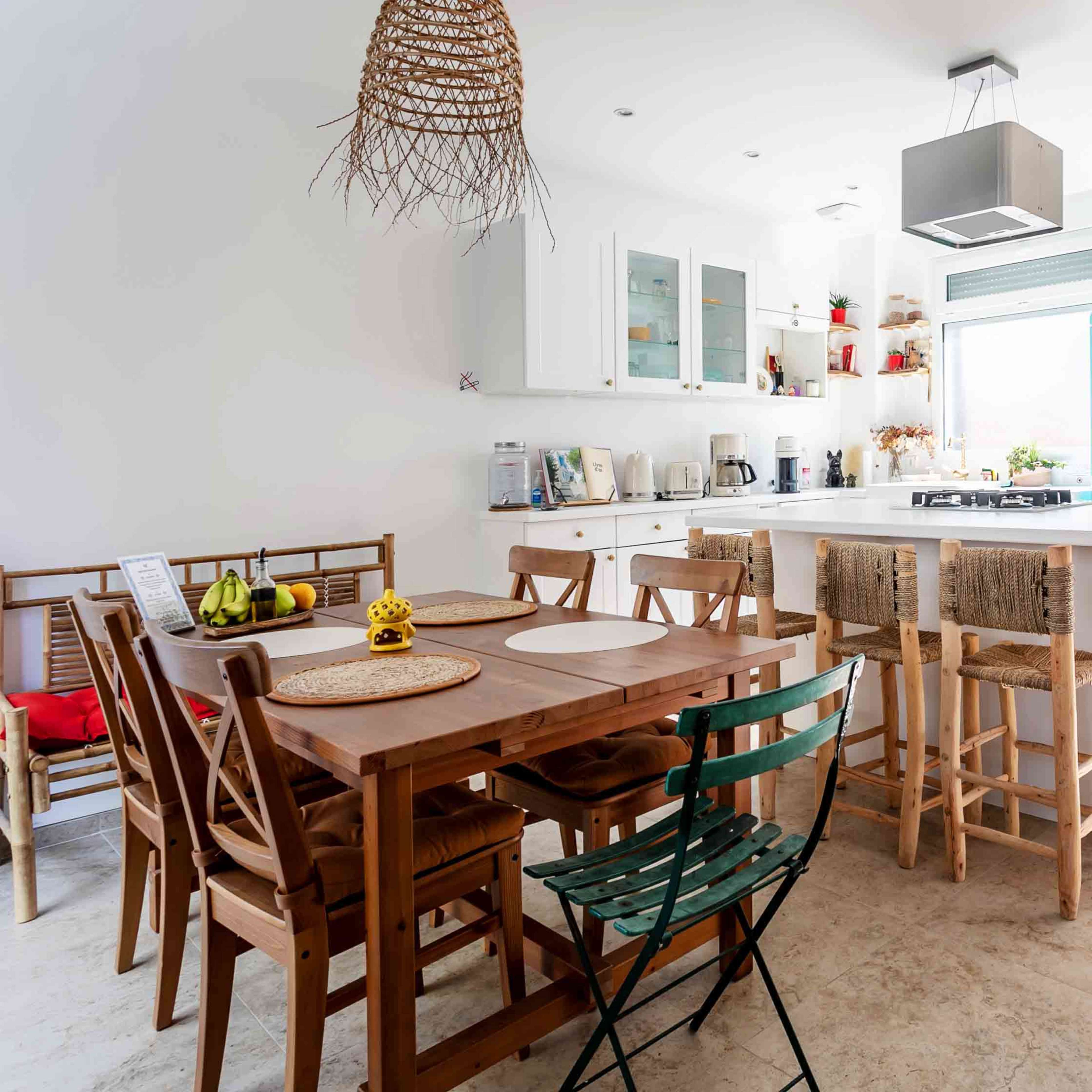 A modern kitchen features a wooden dining table with six chairs, a turquoise refrigerator, and an island with bar stools, alongside a view of a garden through the glass door.