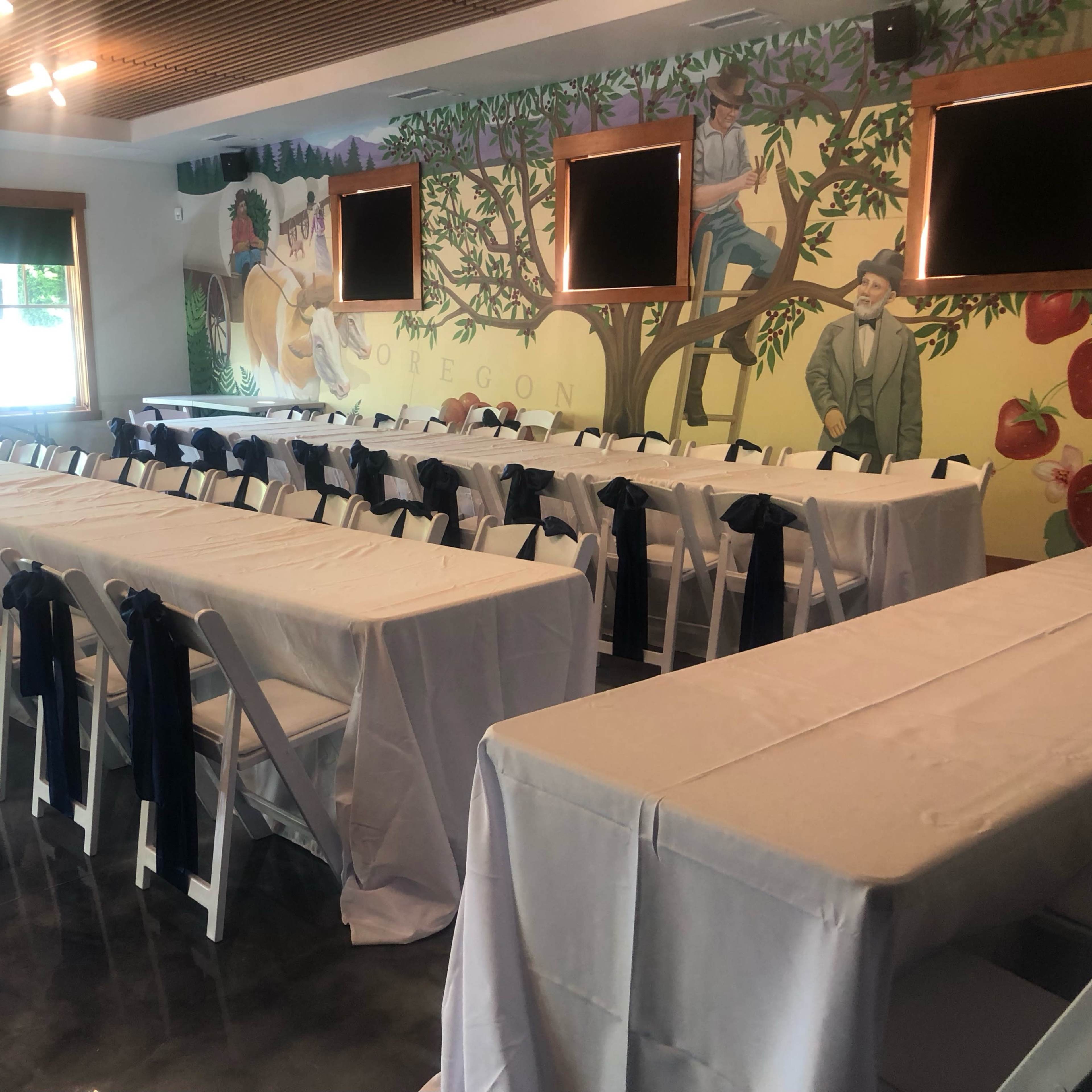 A spacious dining area features rows of tables covered with white tablecloths and adorned with navy blue chair sashes, set against a mural depicting people in a rural scene.