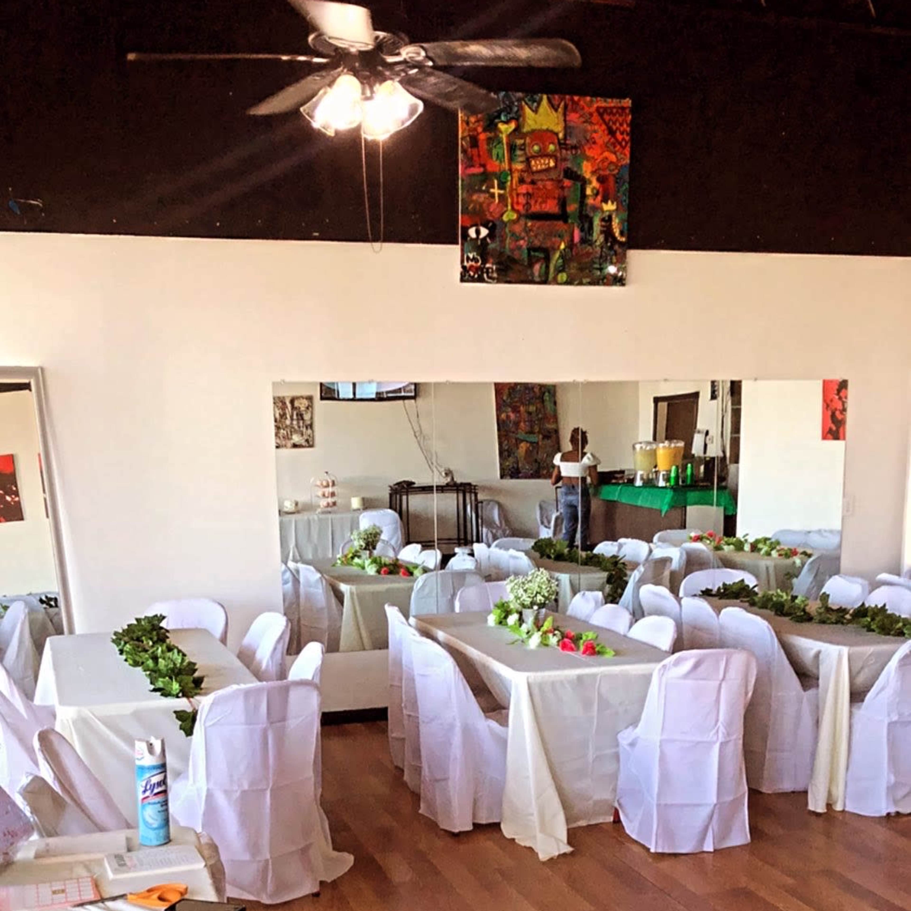 A decorated event space with tables covered in white tablecloths and chairs draped in white fabric, arranged for a gathering.