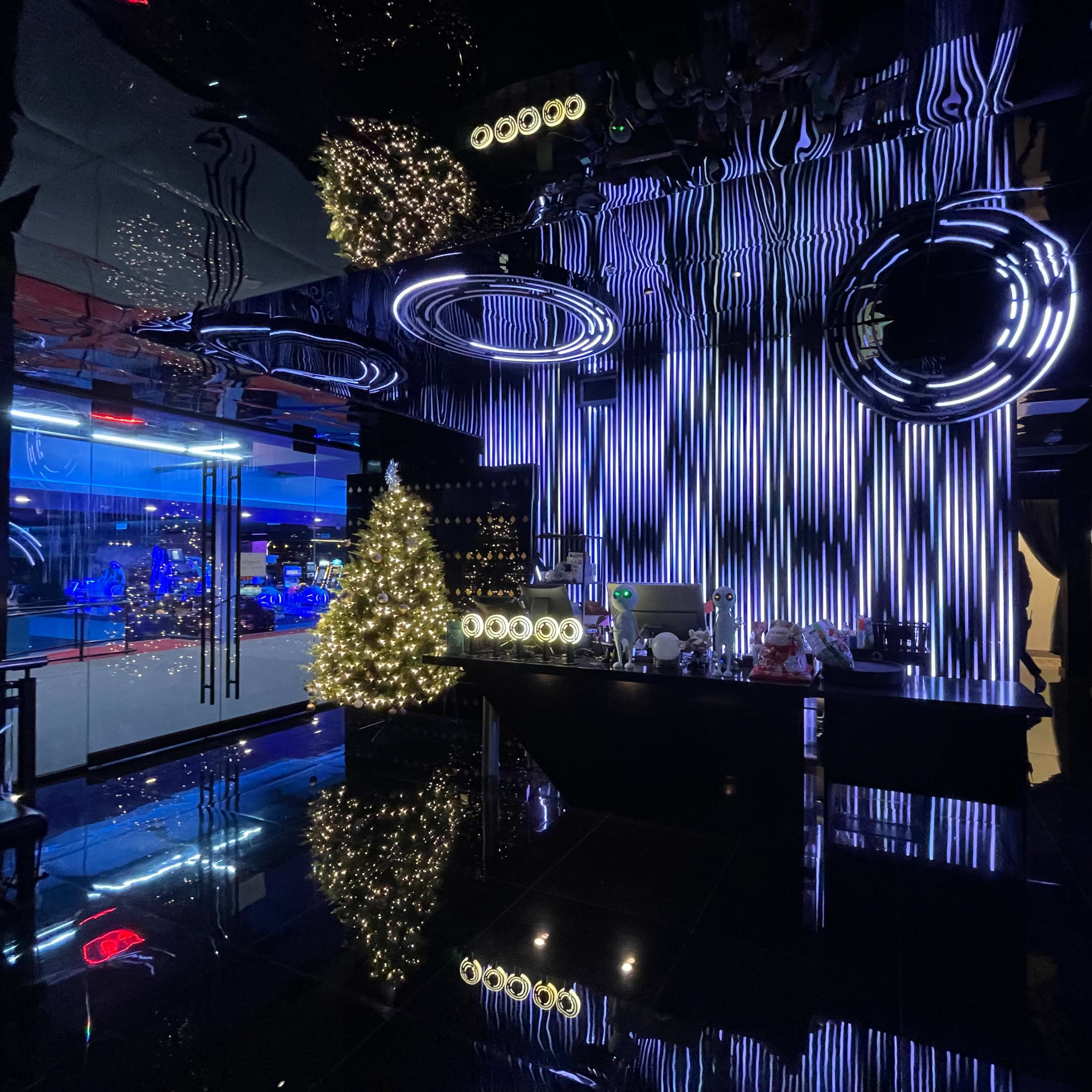 A modern bar environment with a black reflective floor, striking neon lights, and a lit Christmas tree.