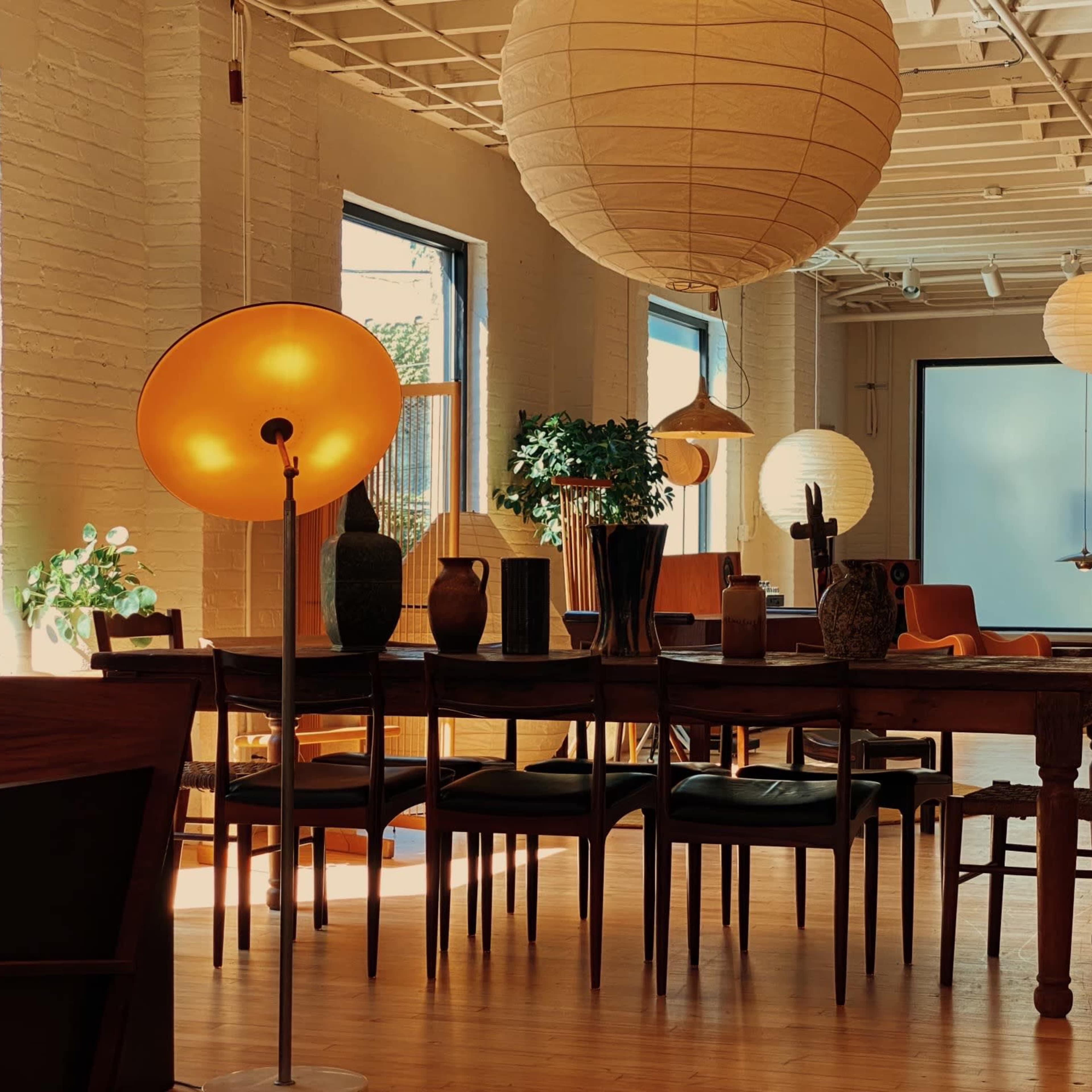 A spacious room with large windows, featuring a wooden dining table surrounded by chairs, various decorative plants, and a mix of modern and vintage lighting fixtures.