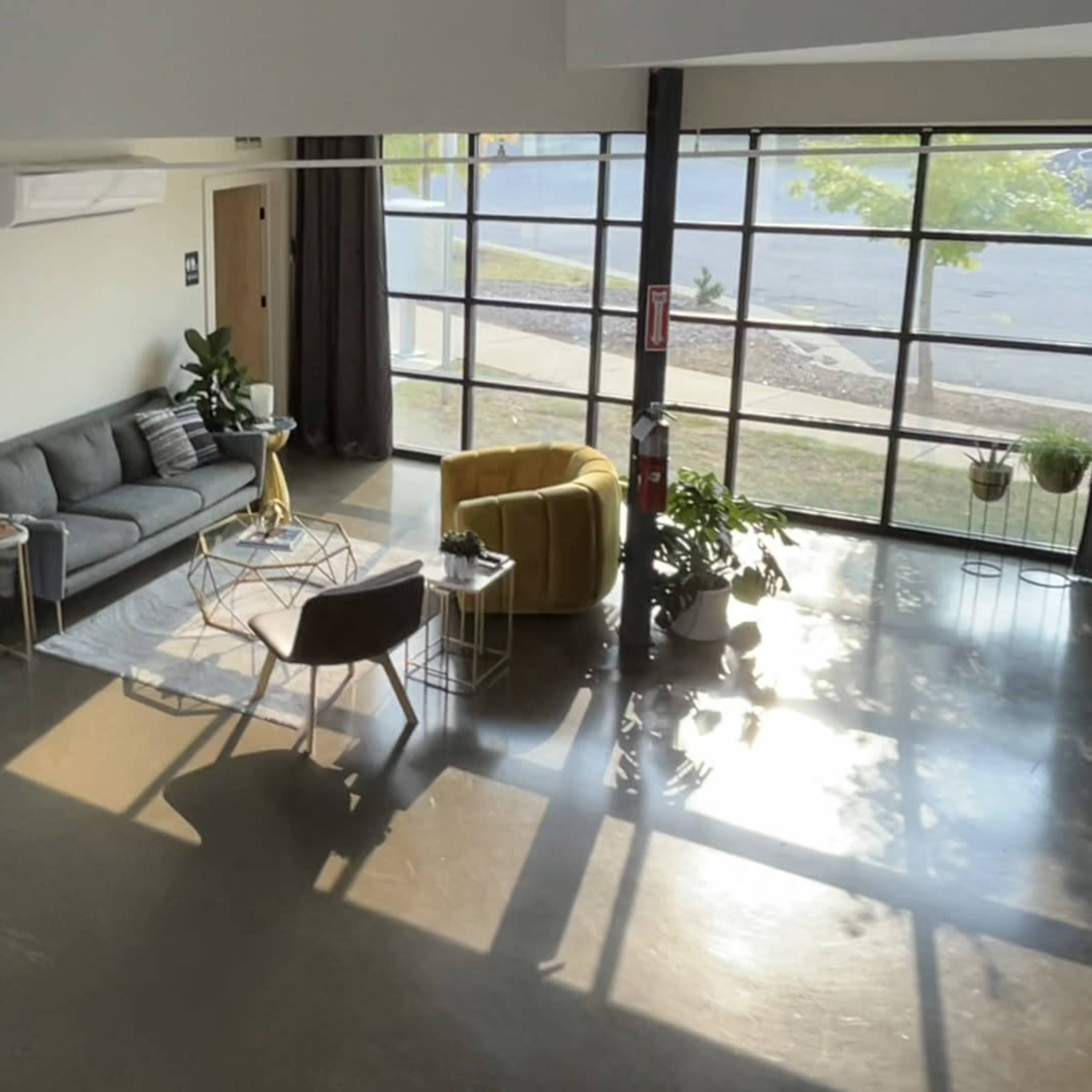 A modern living space features a gray sofa and a yellow chair, surrounded by plants and large windows.