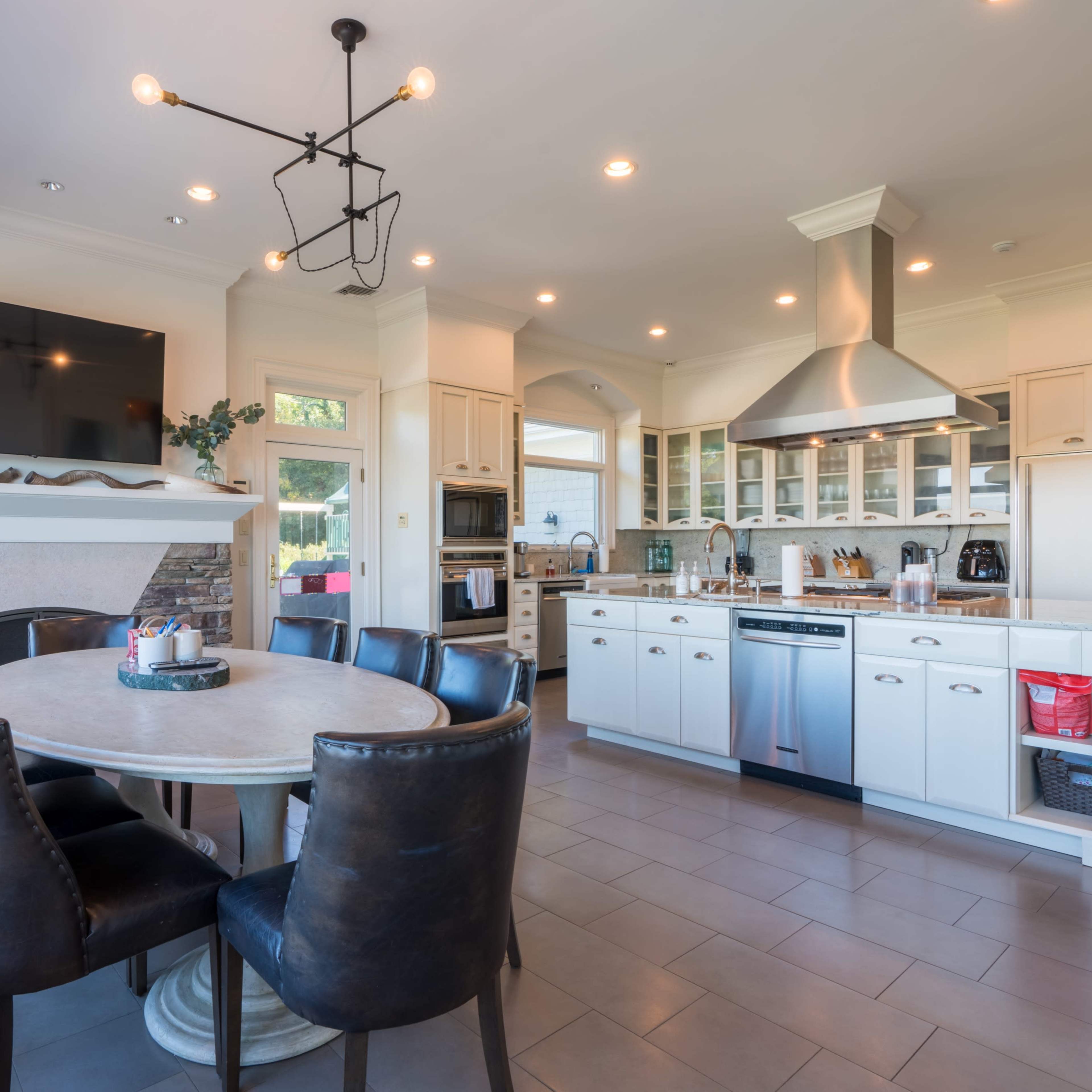 A modern kitchen features a circular dining table surrounded by black chairs, with an island counter, stainless steel appliances, and a stone fireplace.