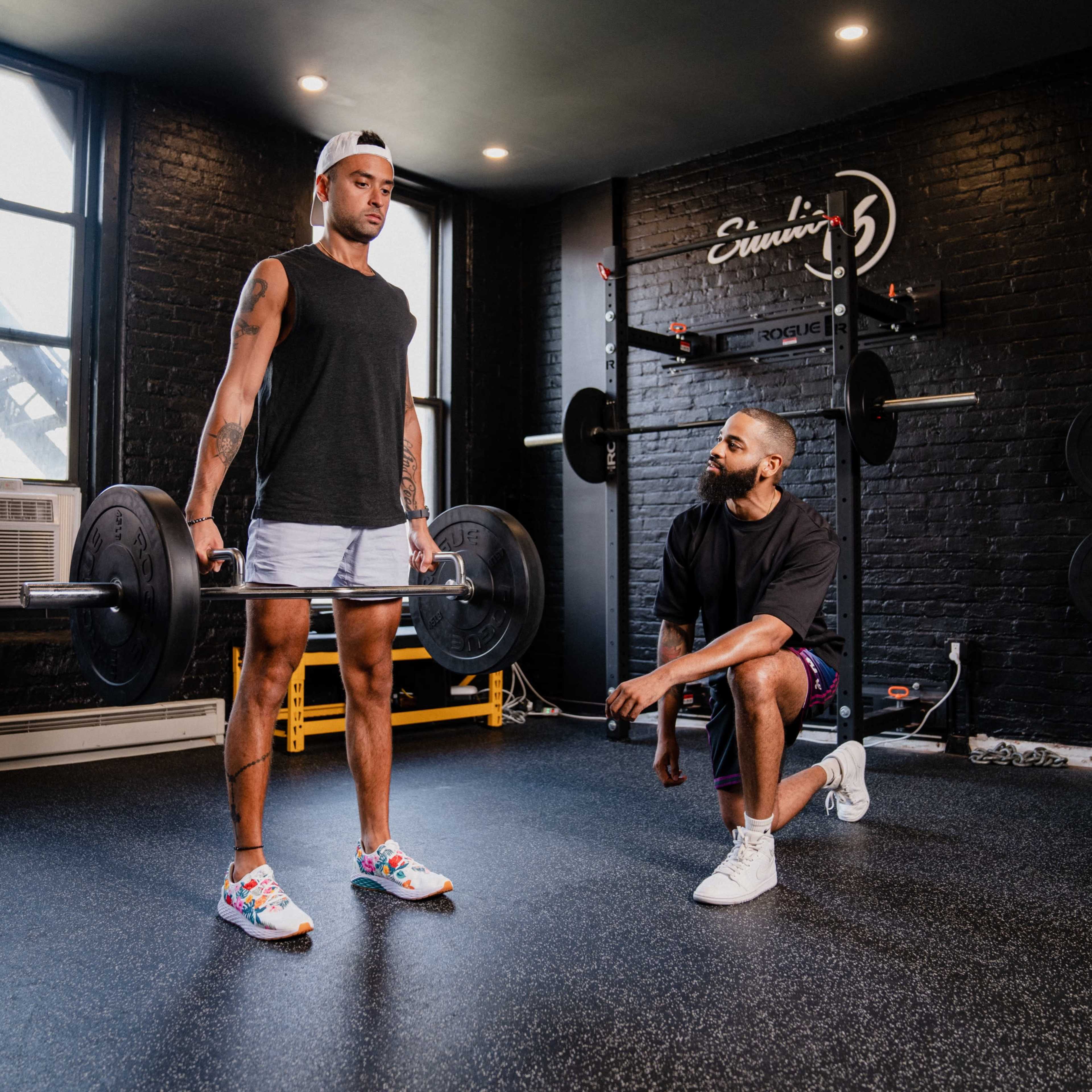 A man performs a deadlift with a barbell while another man kneels beside him, observing in a gym with black walls and equipment in the background.