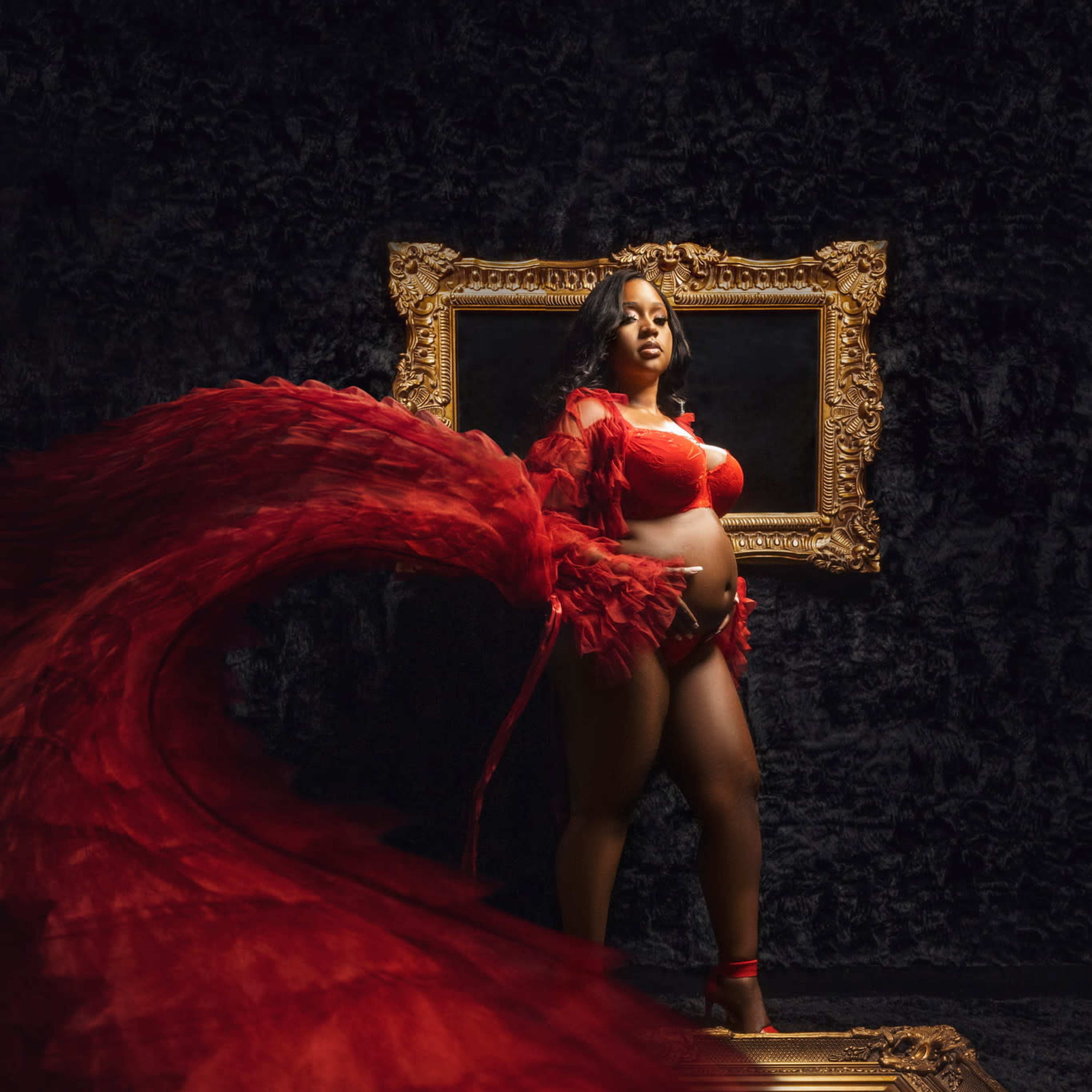 A pregnant woman in a red flowing gown stands before an ornate gold-framed mirror against a dark backdrop.