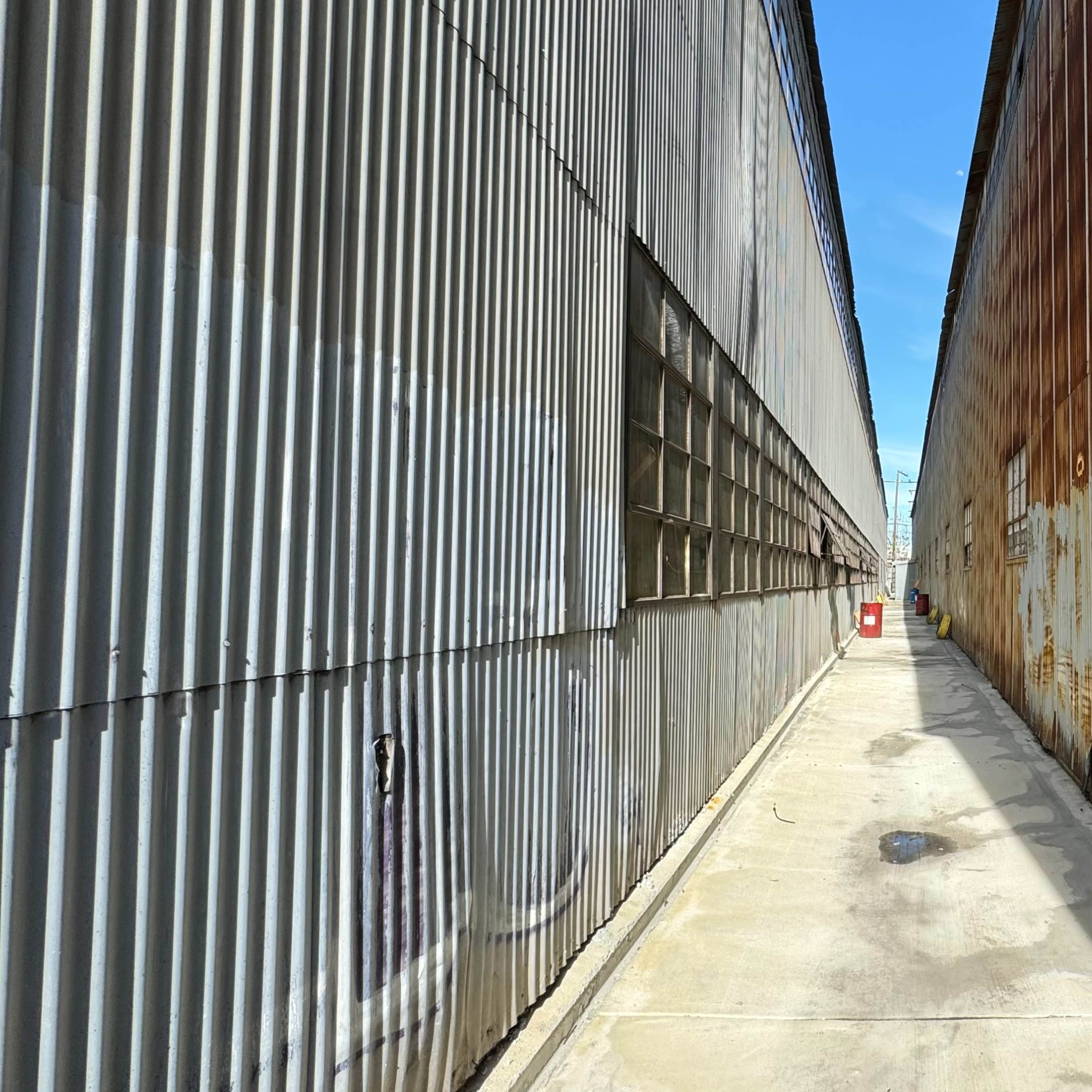 A narrow passageway separates two corrugated metal structures, one weathered and rusty, under a clear blue sky.