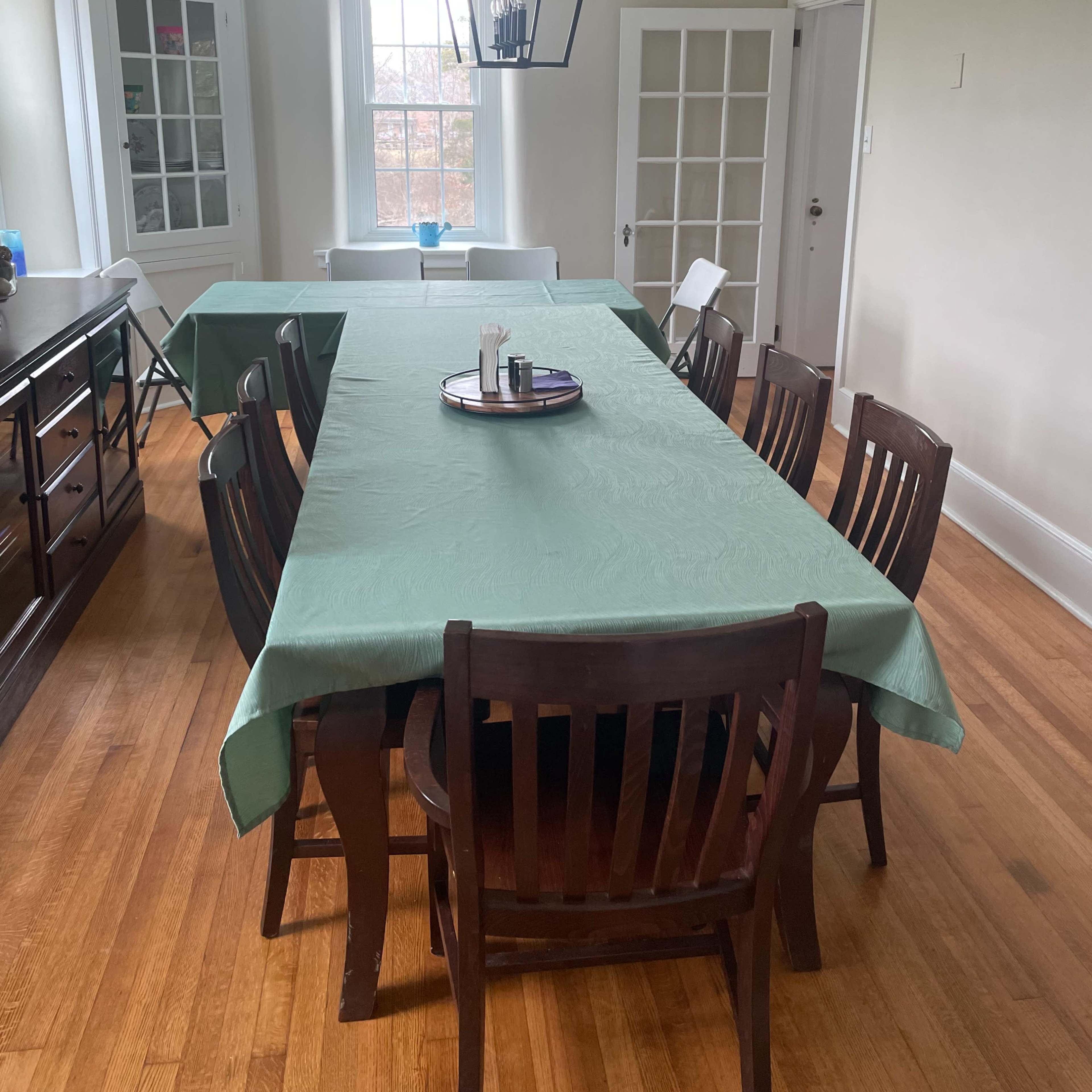 A rectangular dining table with a green tablecloth is set in a bright room featuring light wood floors and a large window.