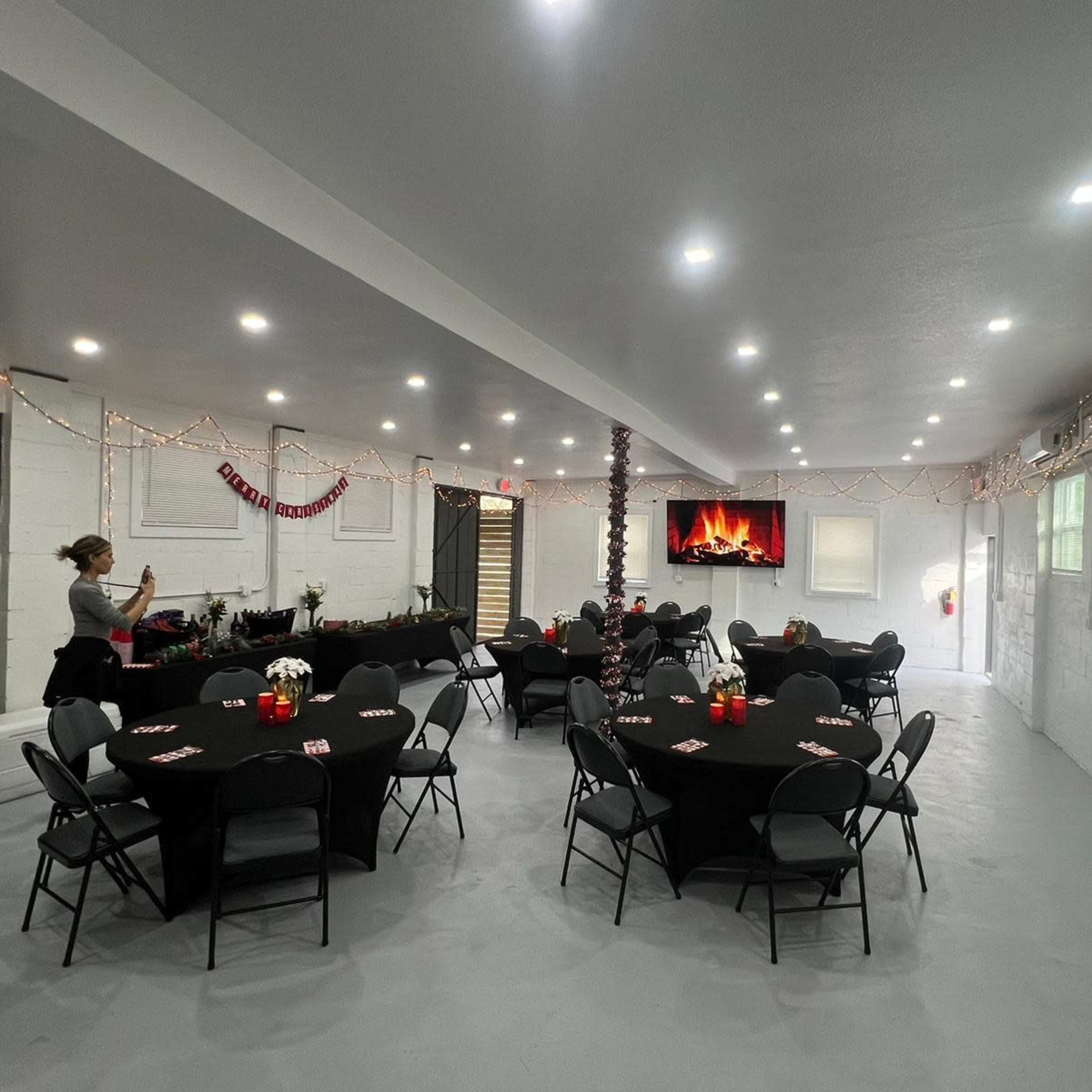 The room features several round tables with black tablecloths, decorated with red centerpieces, and is illuminated by bright overhead lights.
