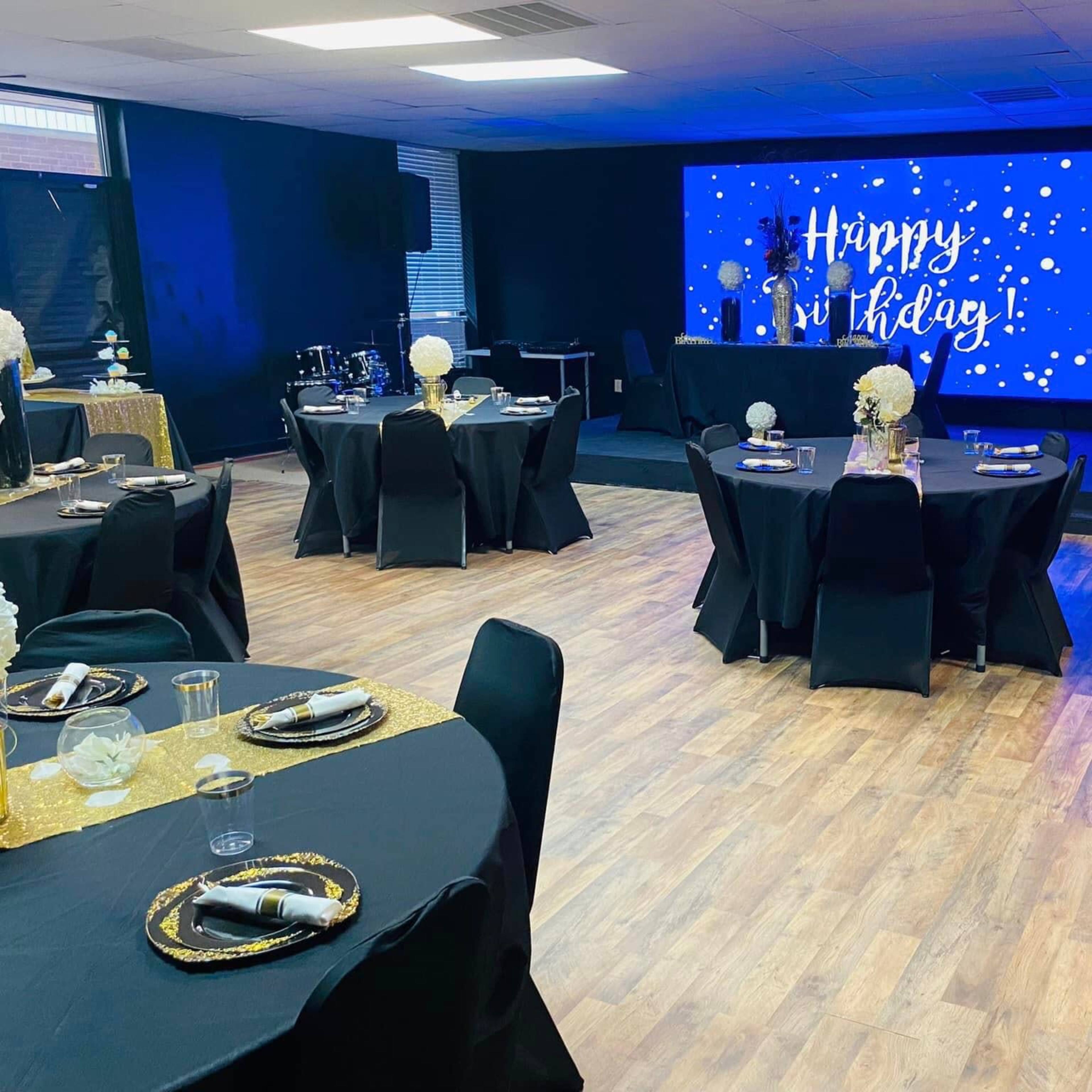 A decorated event space with black tables set for a celebration, a stage featuring a "Happy Birthday" display, and floral arrangements on the tables.