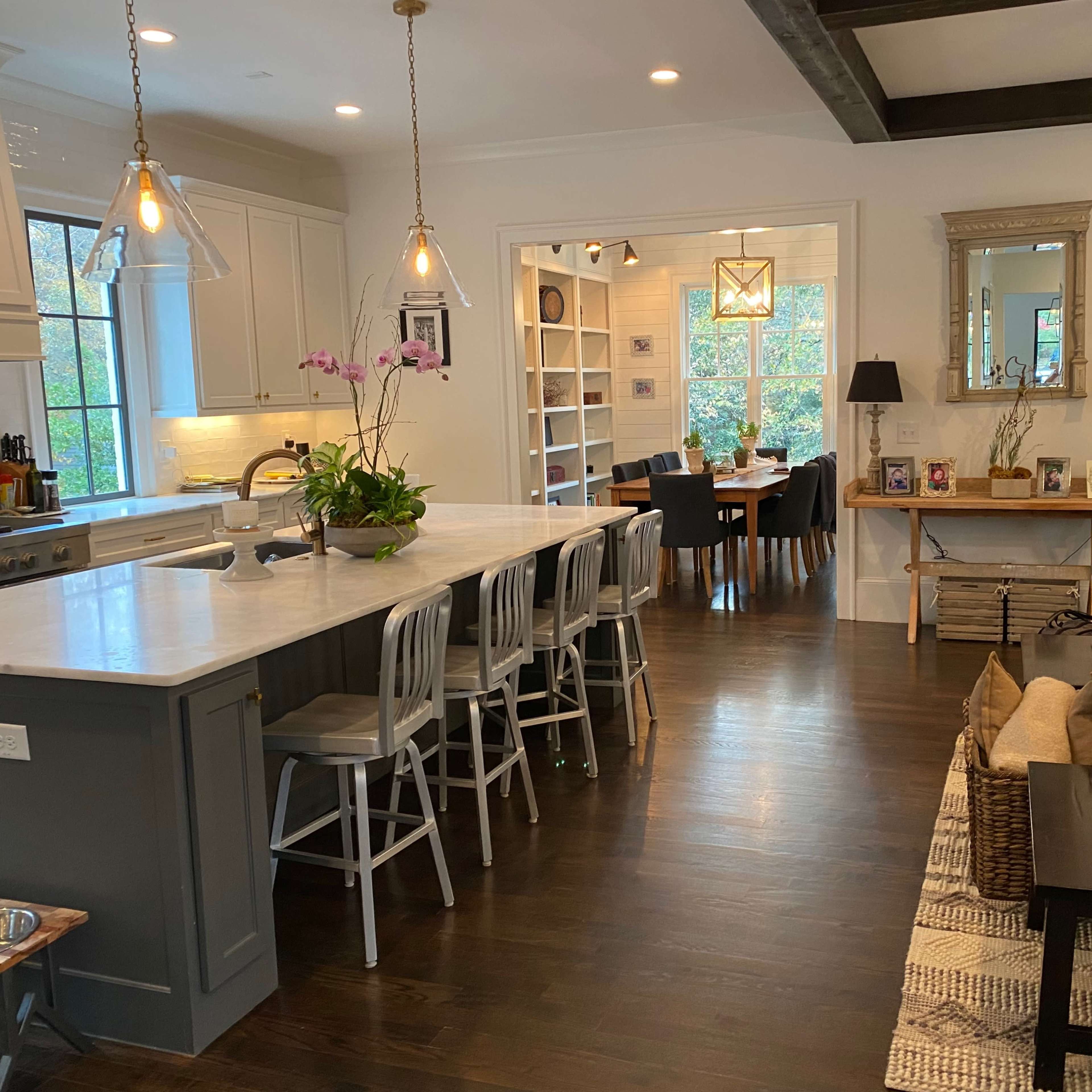 A modern kitchen connected to a dining area, featuring a large island with bar stools, a dining table in the background, and natural light coming through windows.