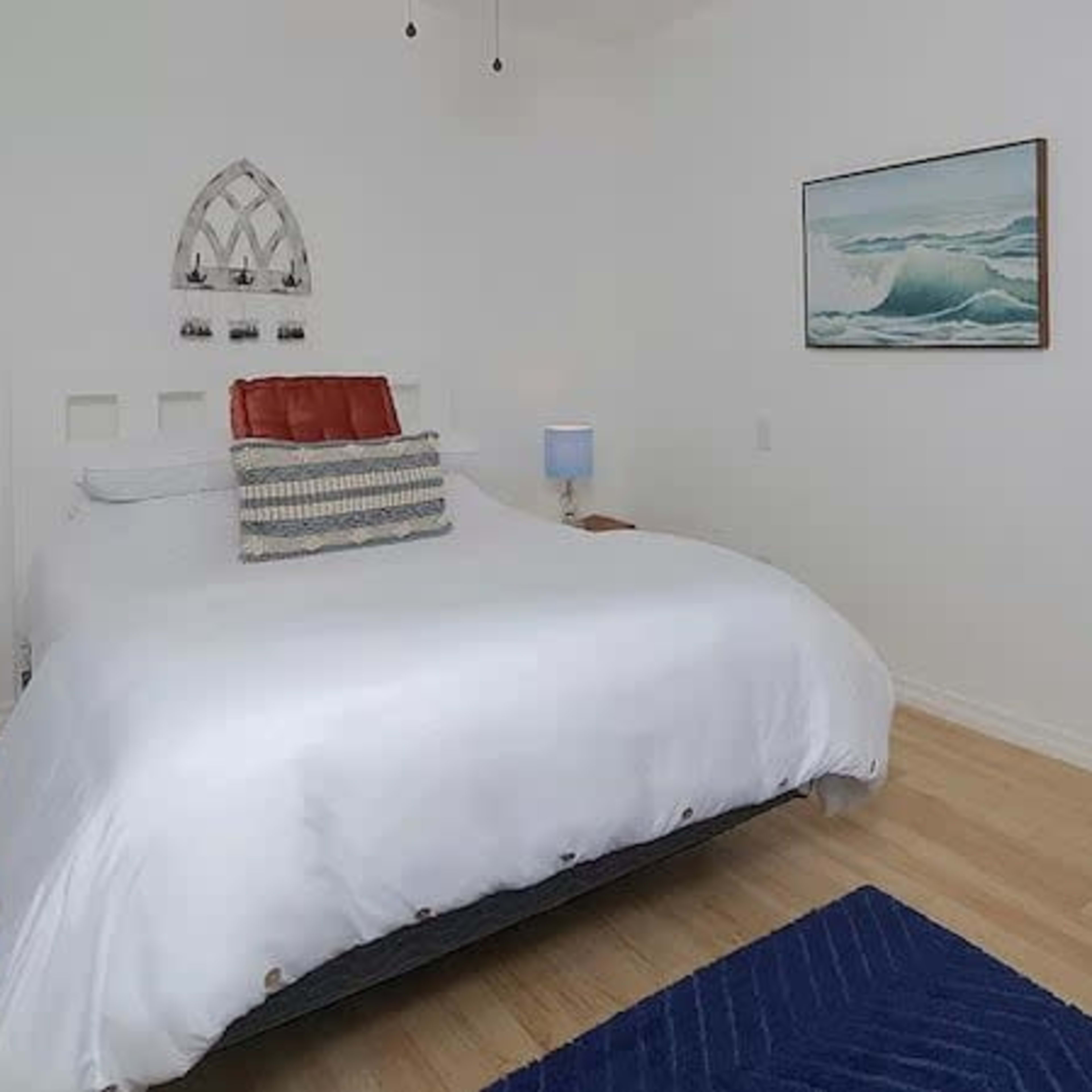 A bright bedroom featuring a large bed with white bedding, two bedside tables with lamps, and a painting of ocean waves on the wall.