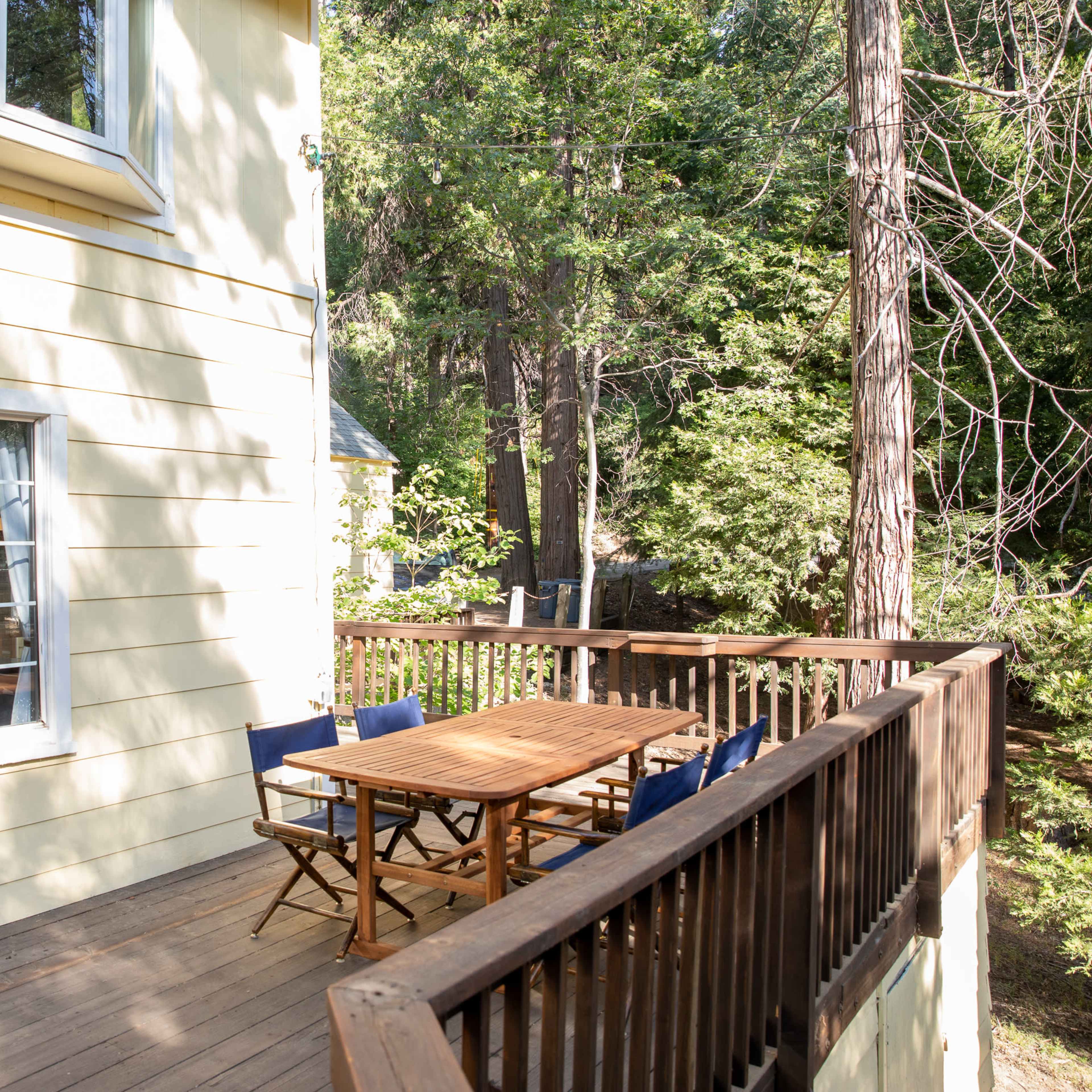 A wooden deck is attached to a yellow house, featuring a large window and a dining table surrounded by chairs.