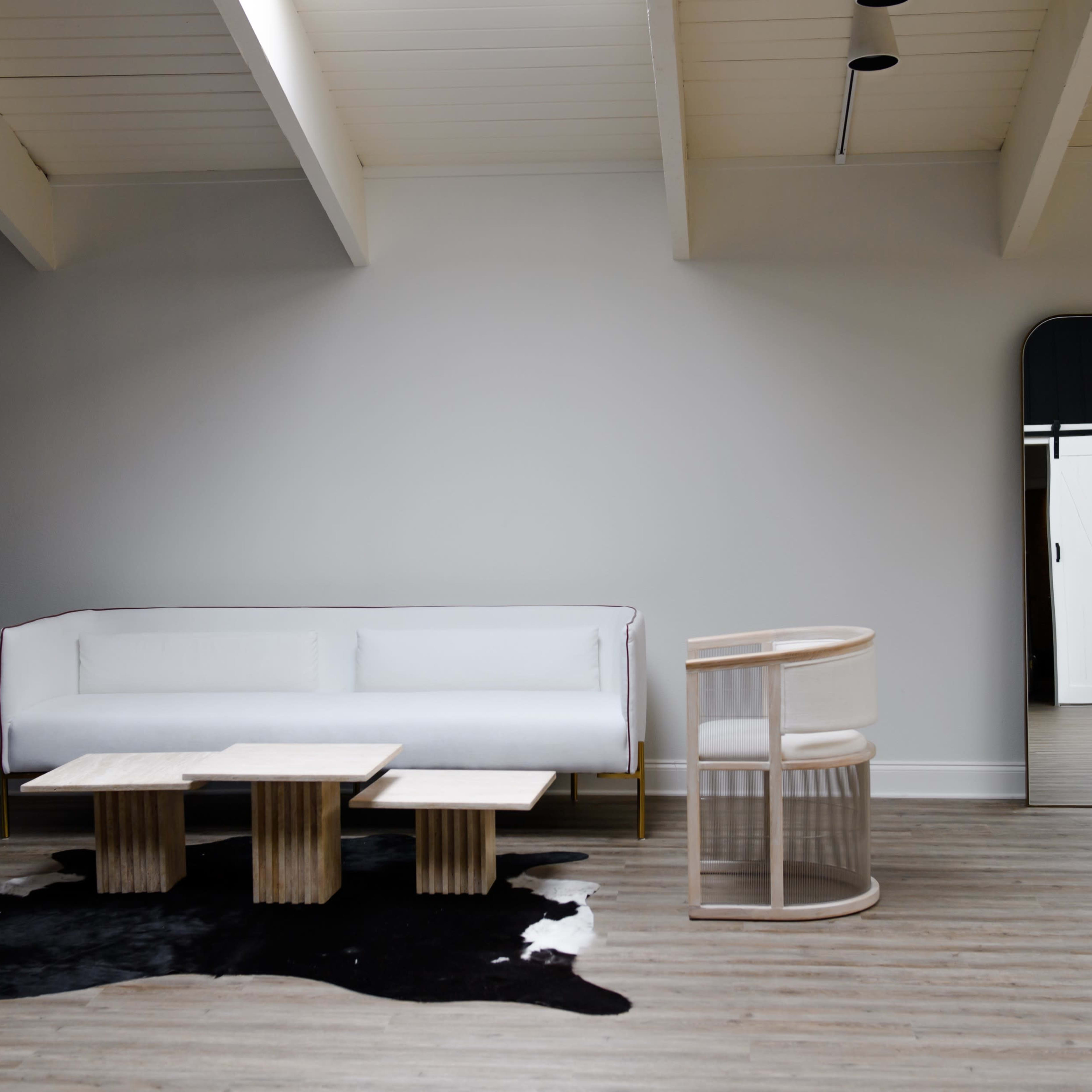A minimalistic living space featuring a white sofa, two modern chairs, and a set of wooden coffee tables arranged on a patterned rug, with a large mirror reflecting a doorway in the background.