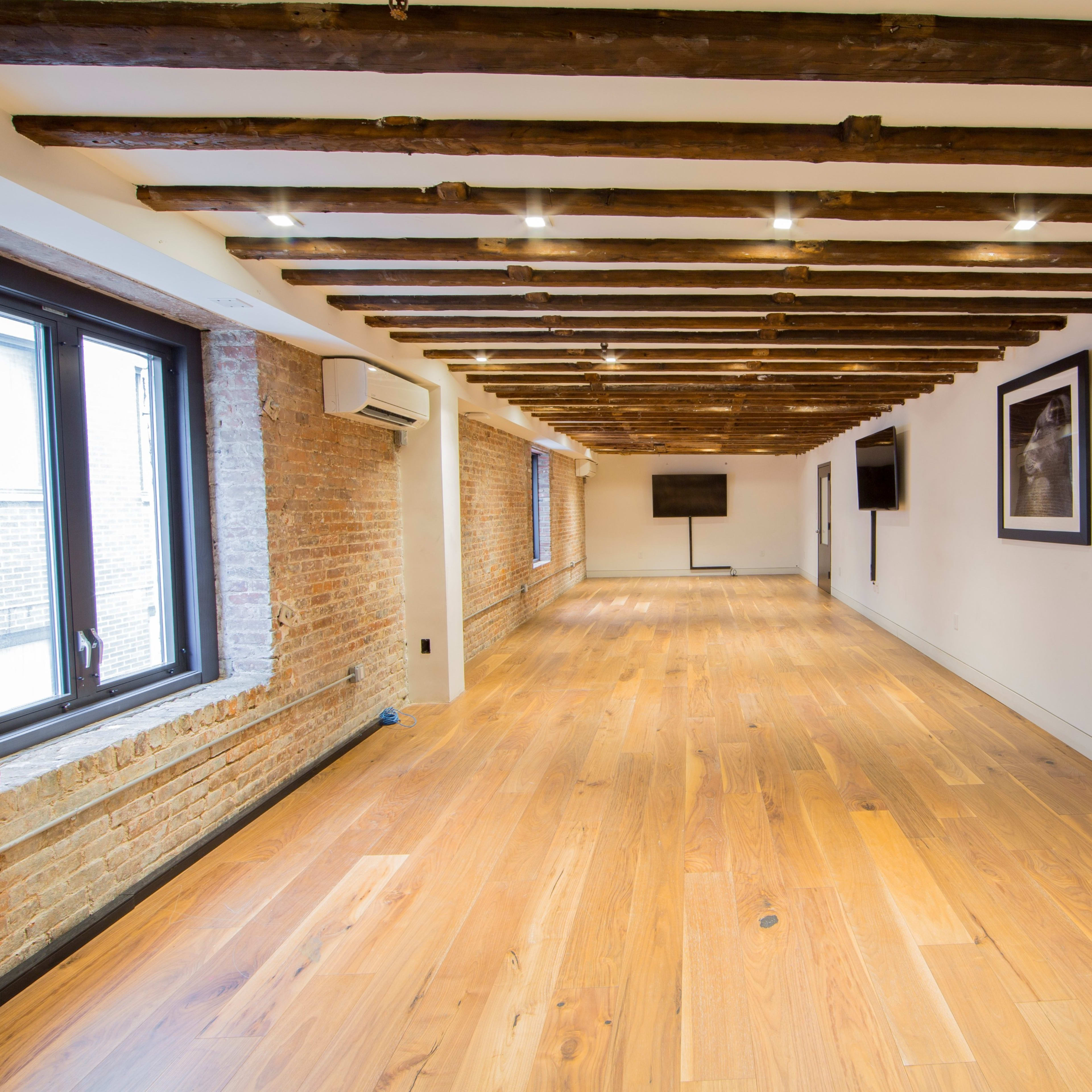 A spacious room with wooden beams on the ceiling, large windows, and hardwood flooring.