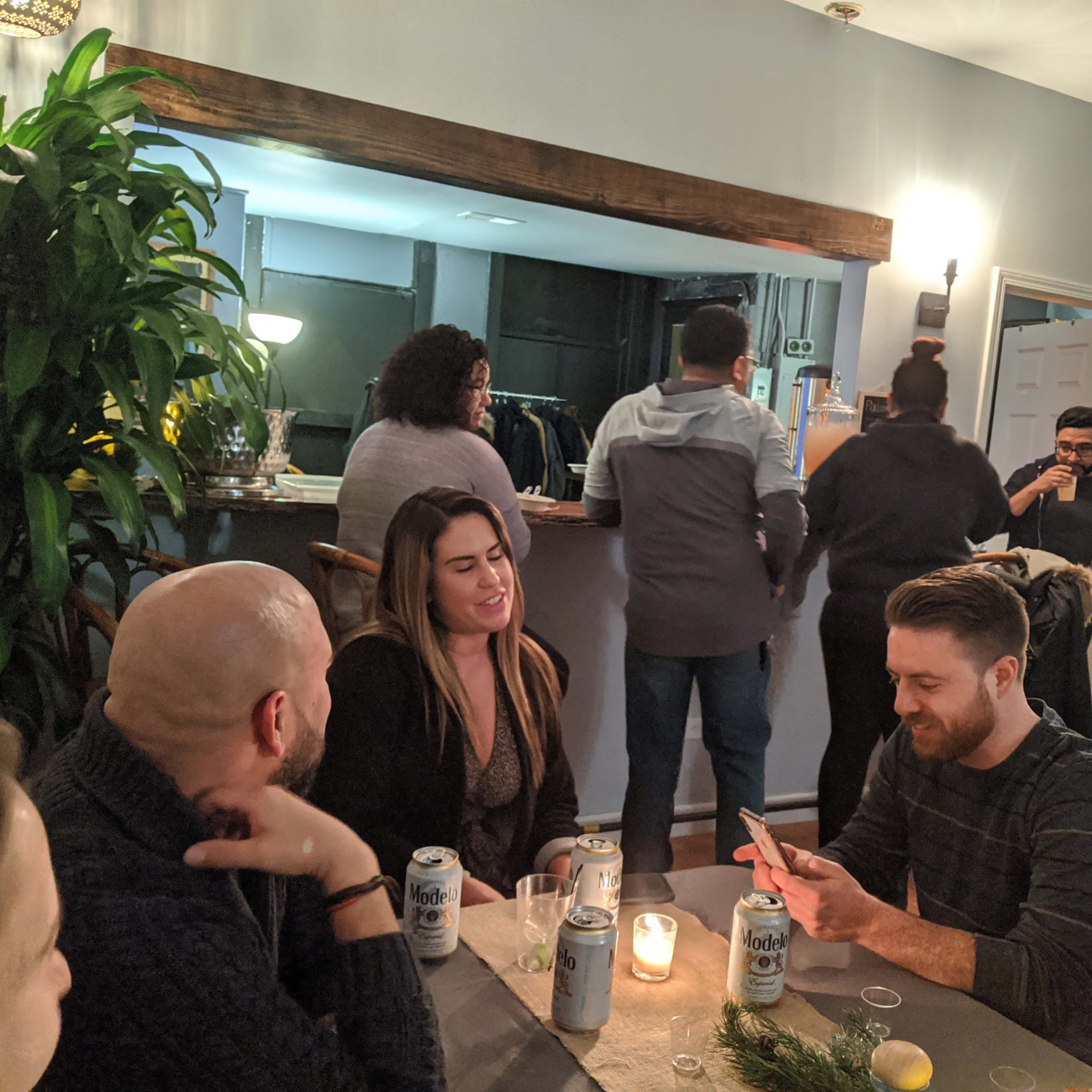 A group of people sit around a table with drinks and snacks while others socialize in the background of a casual indoor gathering.