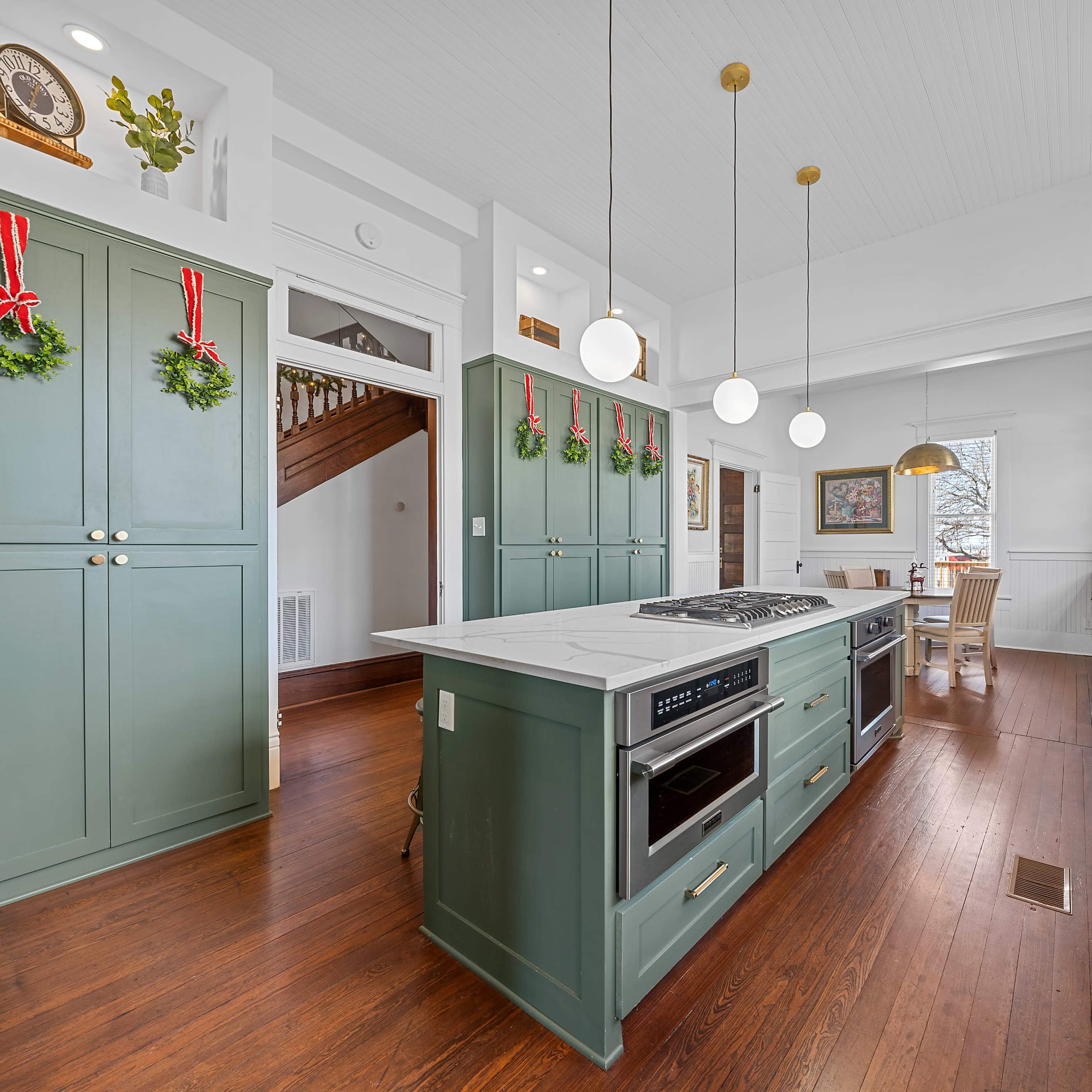 A modern kitchen features green cabinetry, a central island with a stovetop, and large windows providing natural light.