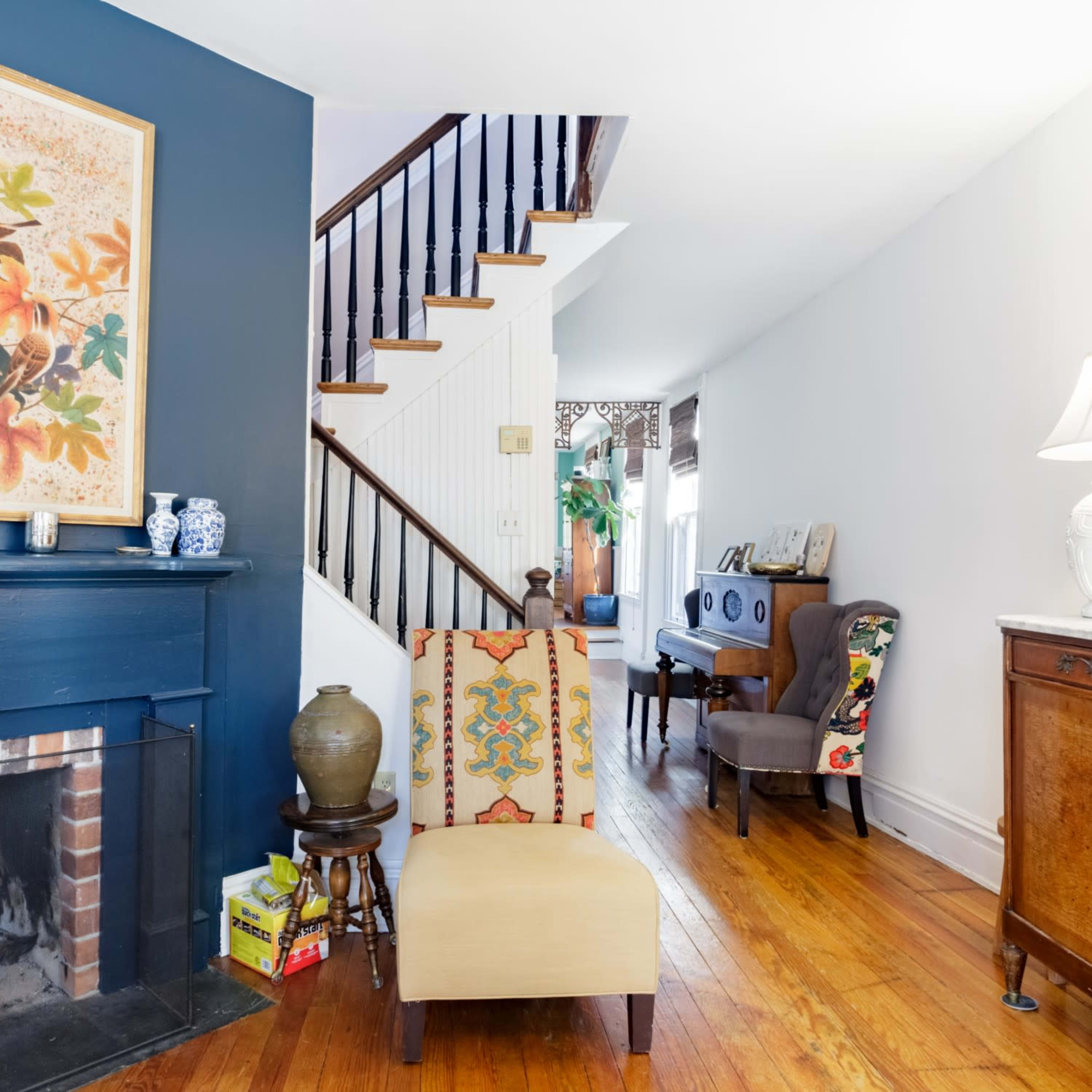 A living room with a fireplace, a colorful flower painting on the blue wall, a staircase in the background, and an arrangement of furniture including a chair and decorative items.