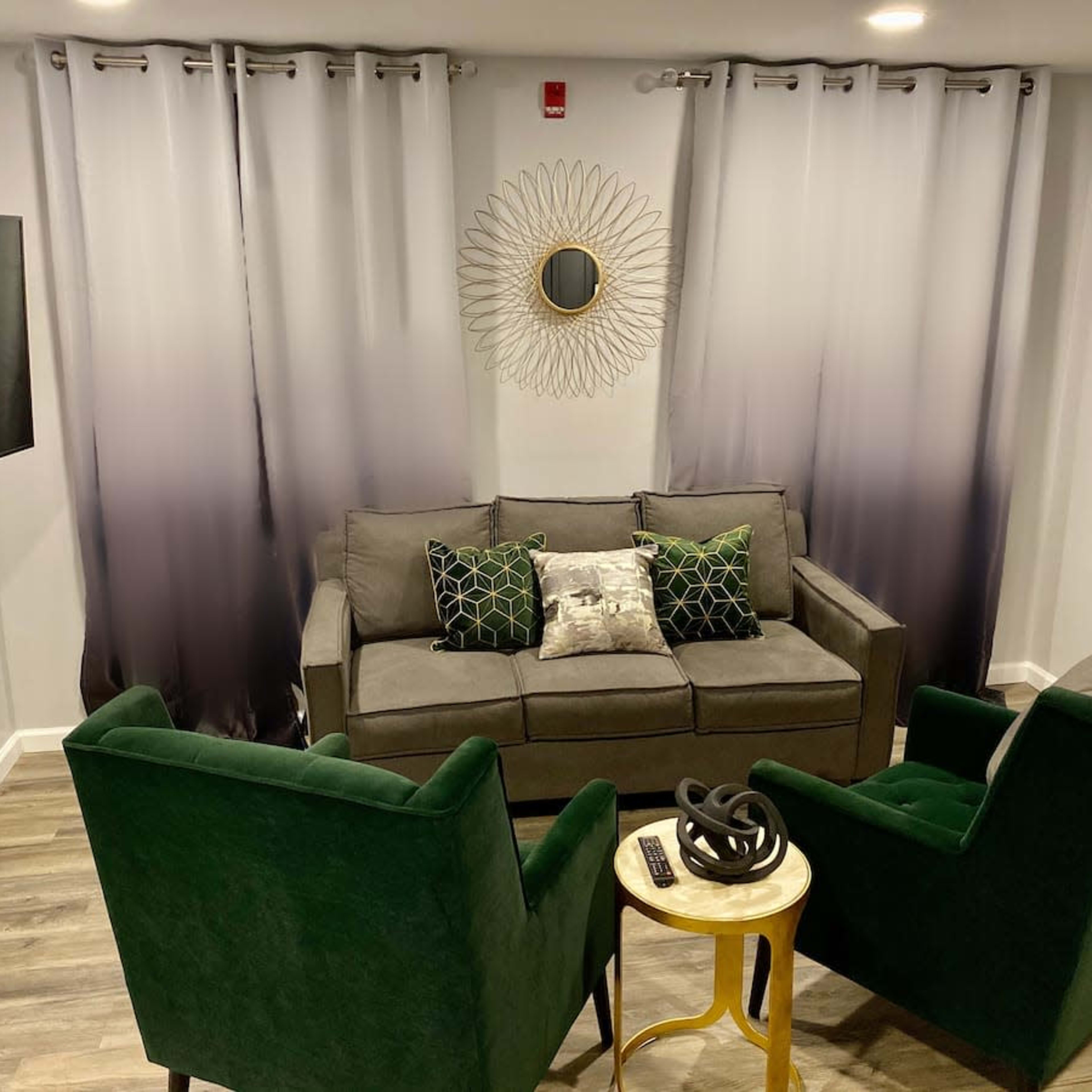 A modern living room featuring a gray sofa with decorative pillows, two green armchairs, a round wall mirror, and a small gold side table.
