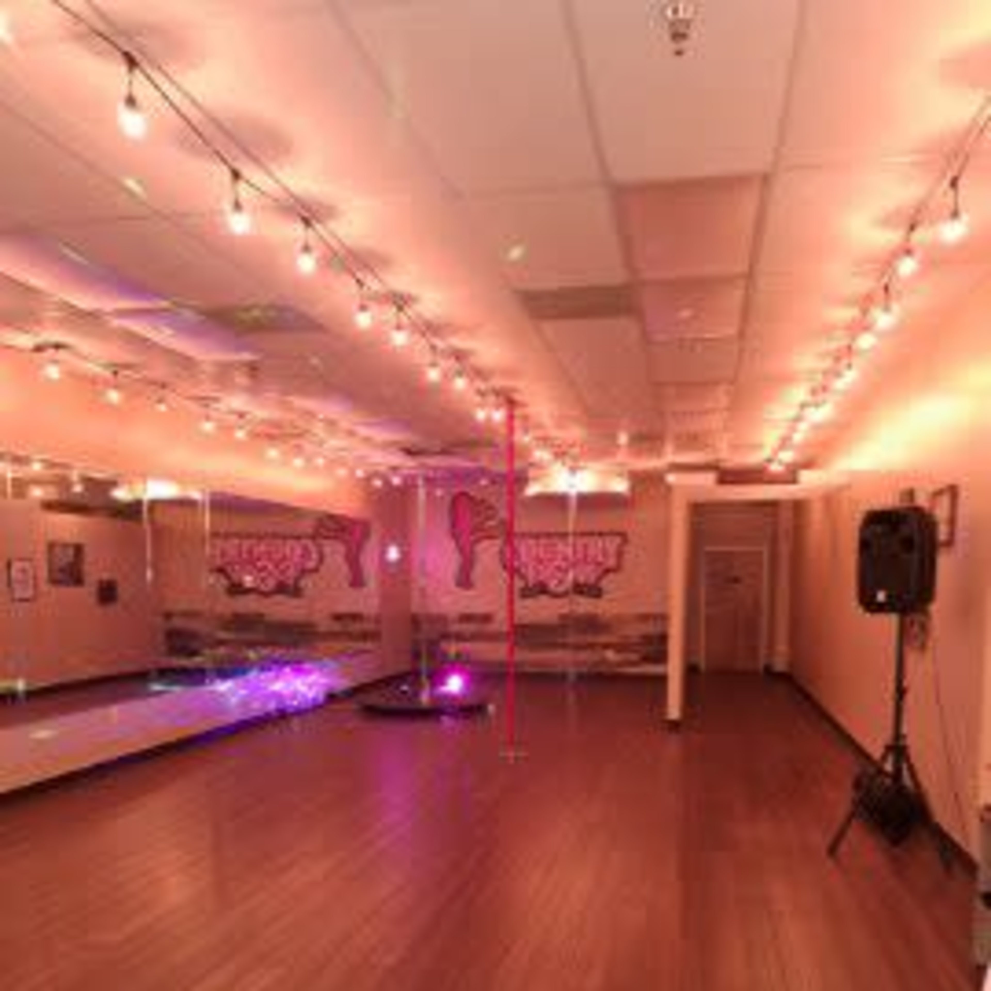 A dance studio with wooden flooring, mirrored walls, and a centrally placed pole, illuminated by string lights and colored spotlights.