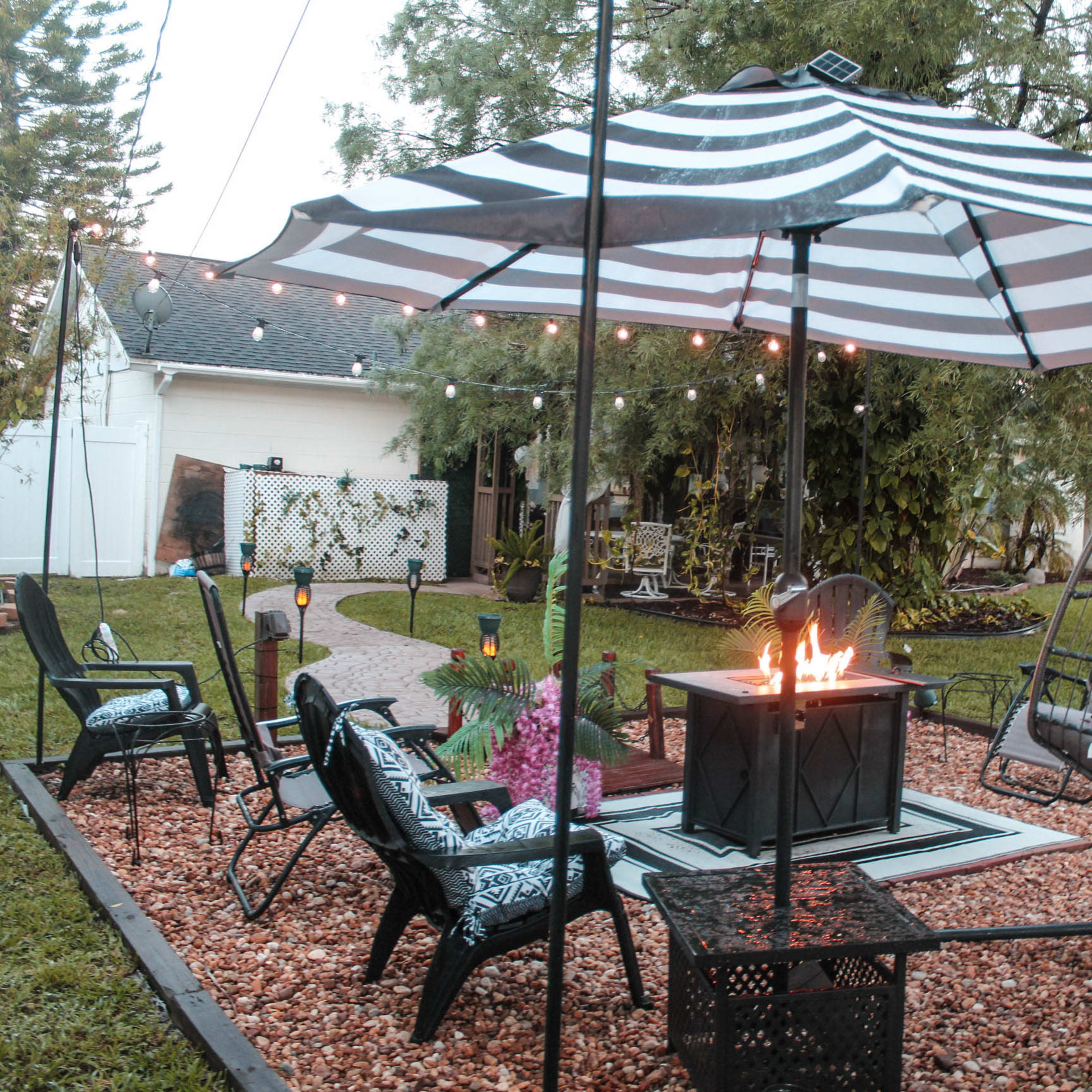 A patio area features a striped umbrella over a fire pit, surrounded by black chairs and a swinging lounge chair, with landscaping and string lights in the background.