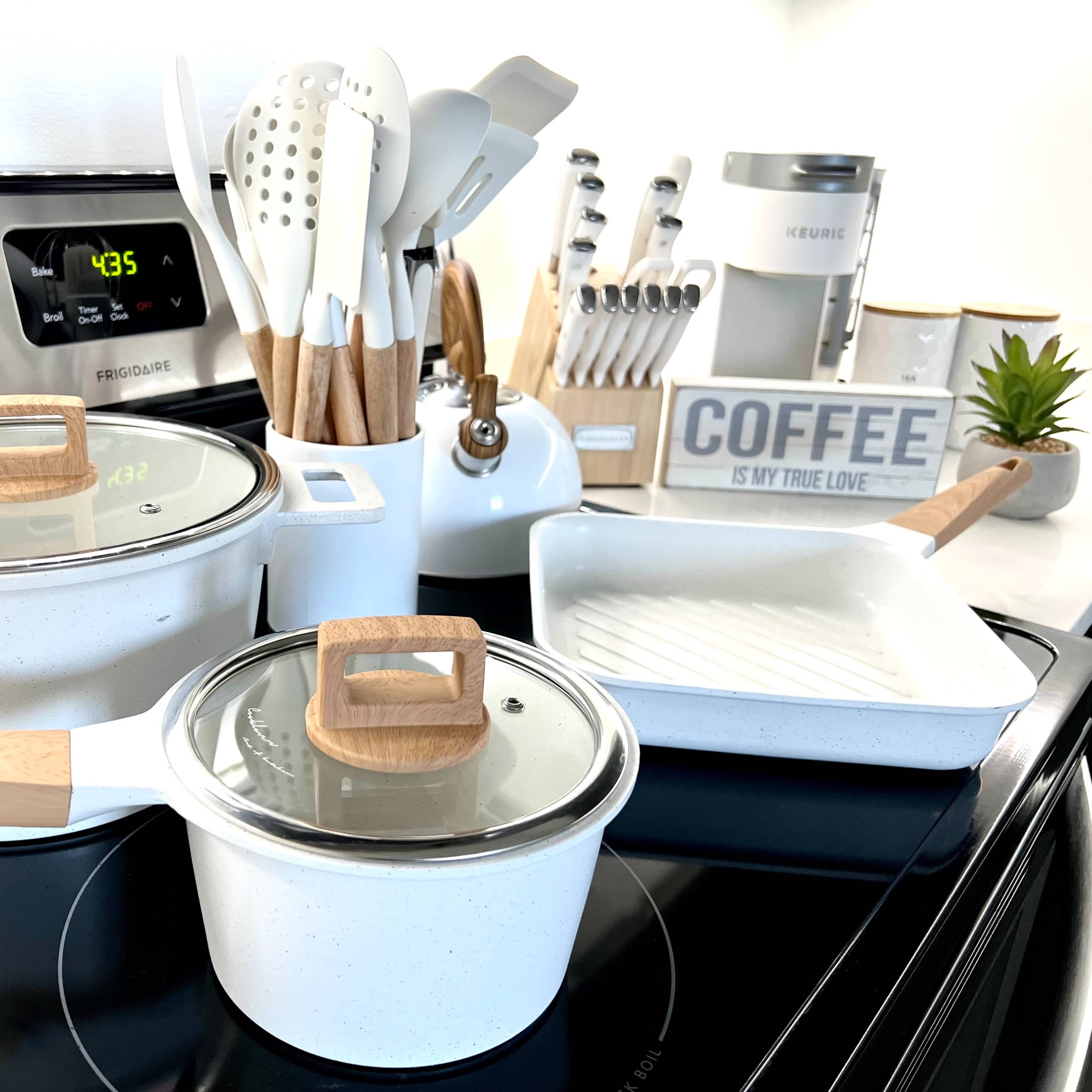 A set of white cooking pots with wooden handles and a glass lid sits on a stove, alongside various kitchen utensils and a coffee-themed decor item.