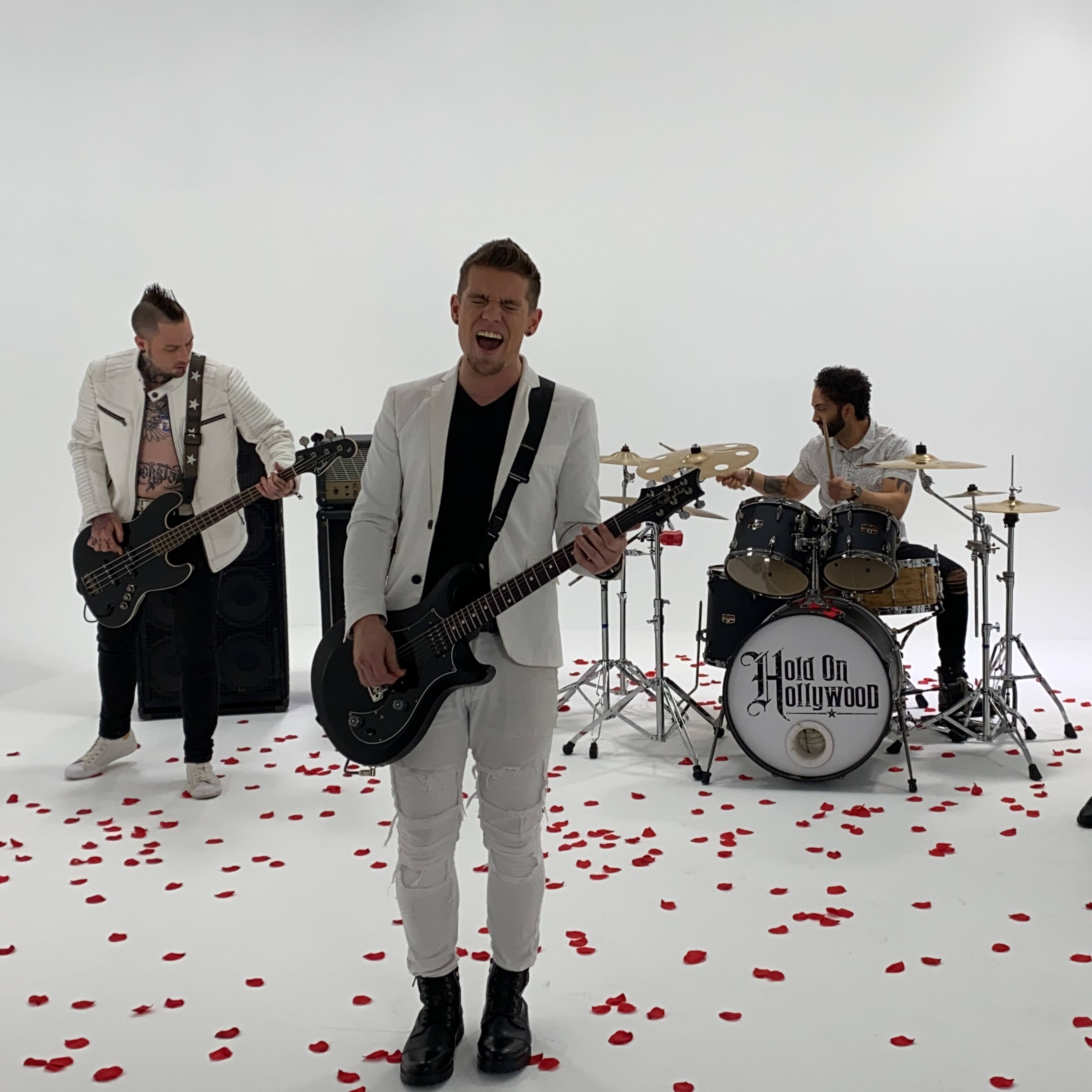 A four-member band is performing on a white stage scattered with rose petals, with one member singing and playing guitar at the front.