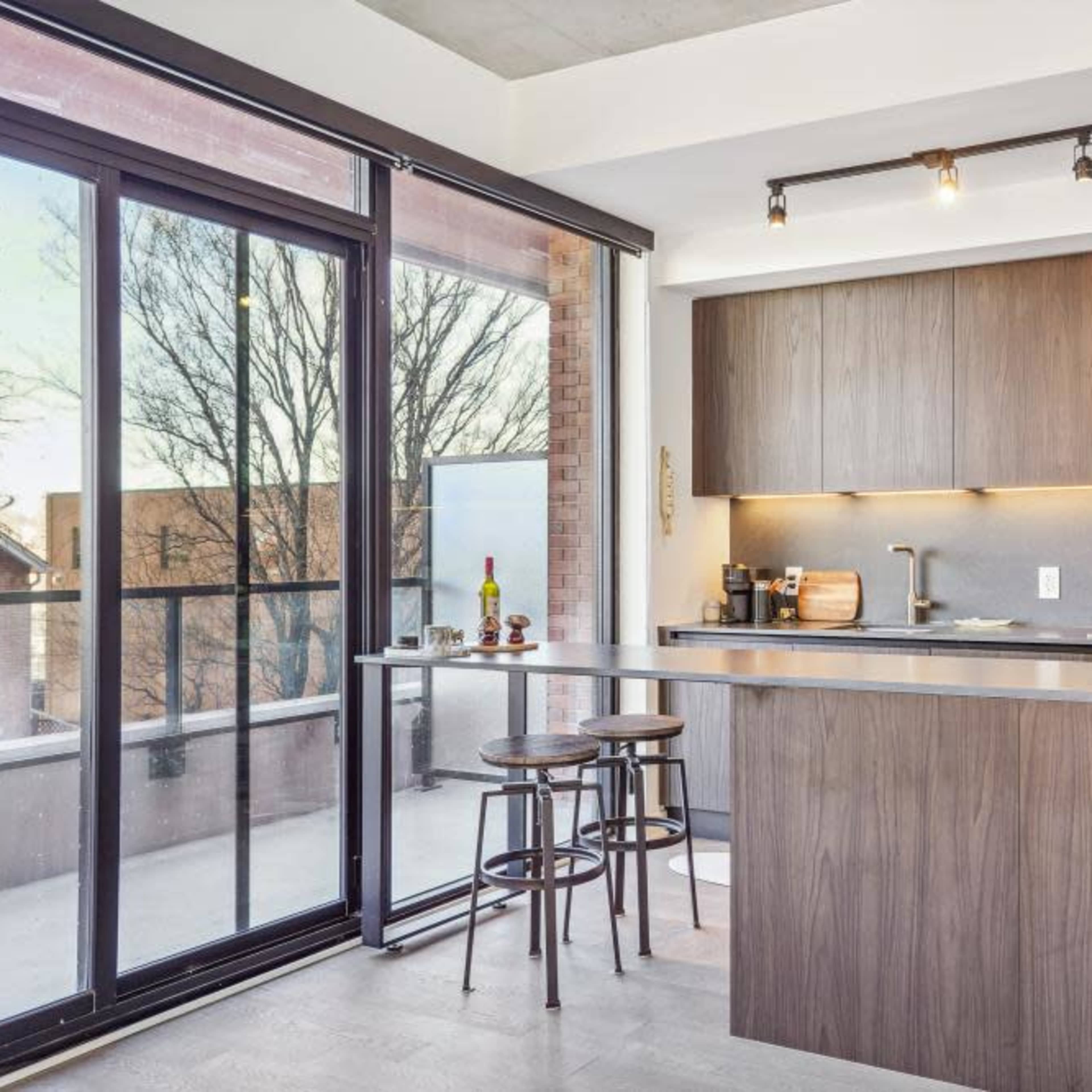 A modern kitchen features a wooden island, bar stools, and large glass doors opening to a balcony with trees visible outside.