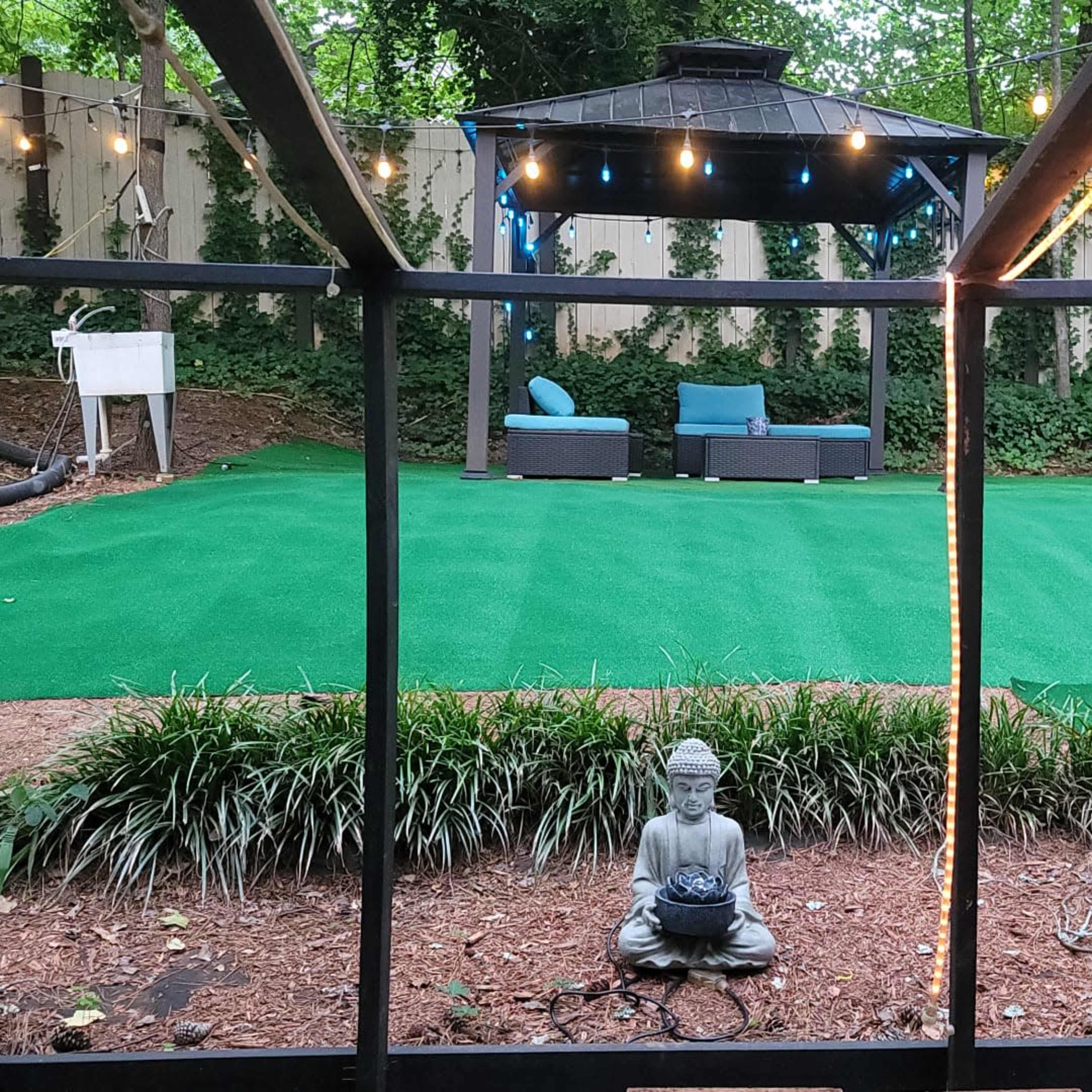 A backyard features an artificial turf area with a gazebo, seating arrangement, and a statue of Buddha surrounded by greenery.
