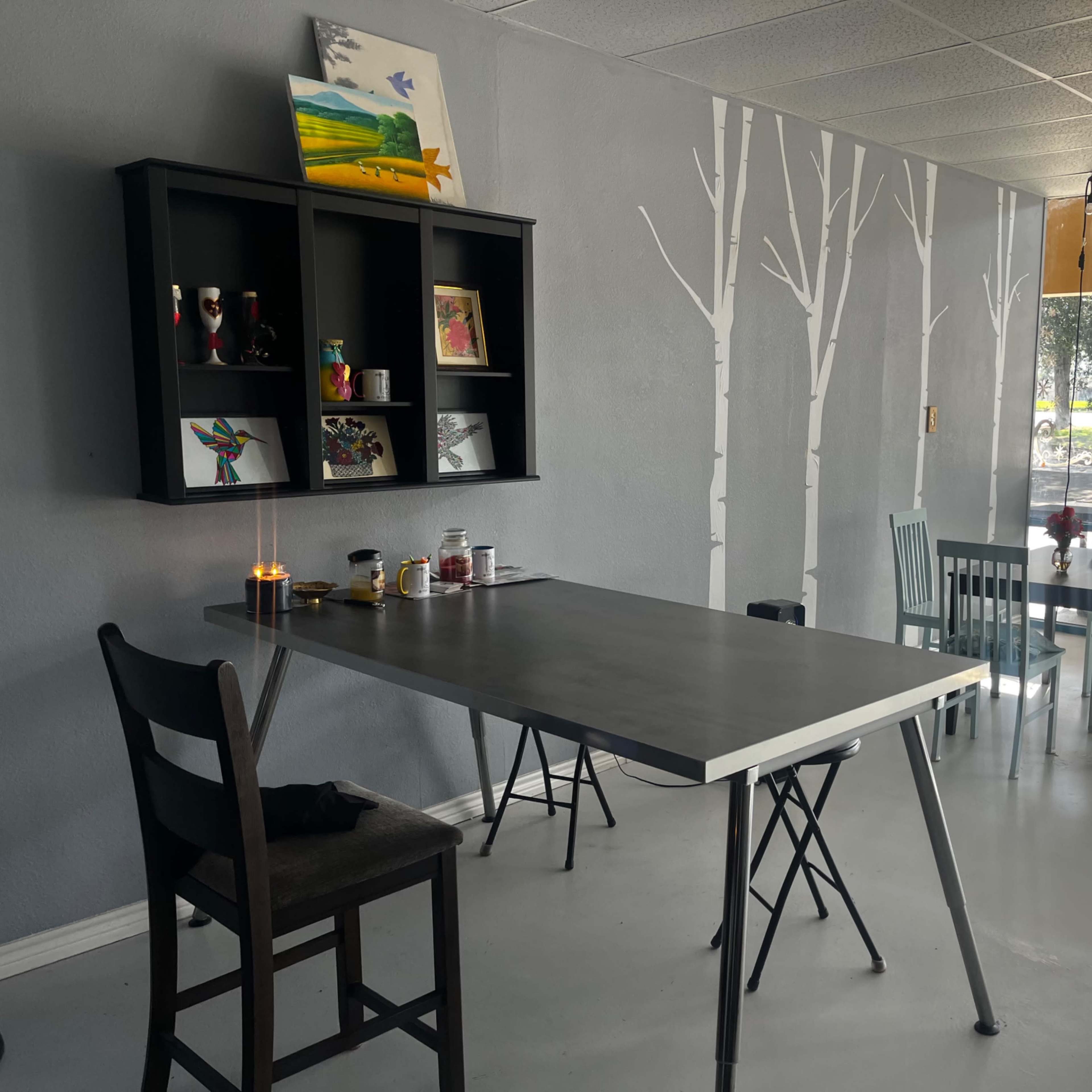 A minimalist café interior featuring a black table with folding legs, a shelf with decorative items, and white tree murals on the gray walls.