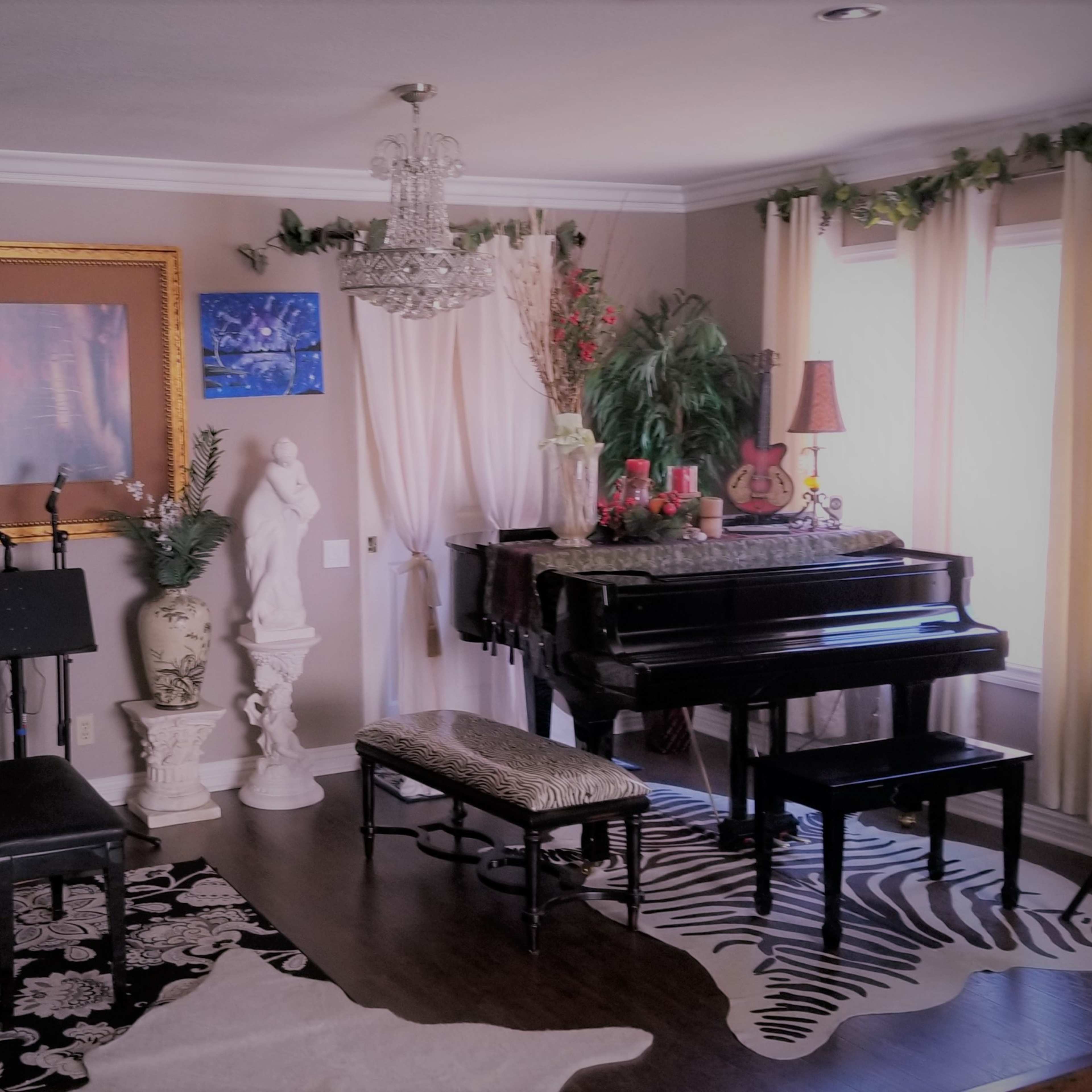 A music room with a grand piano, a small bench, a speaker, and decorative plants, all set against a patterned rug and an elegant chandelier.