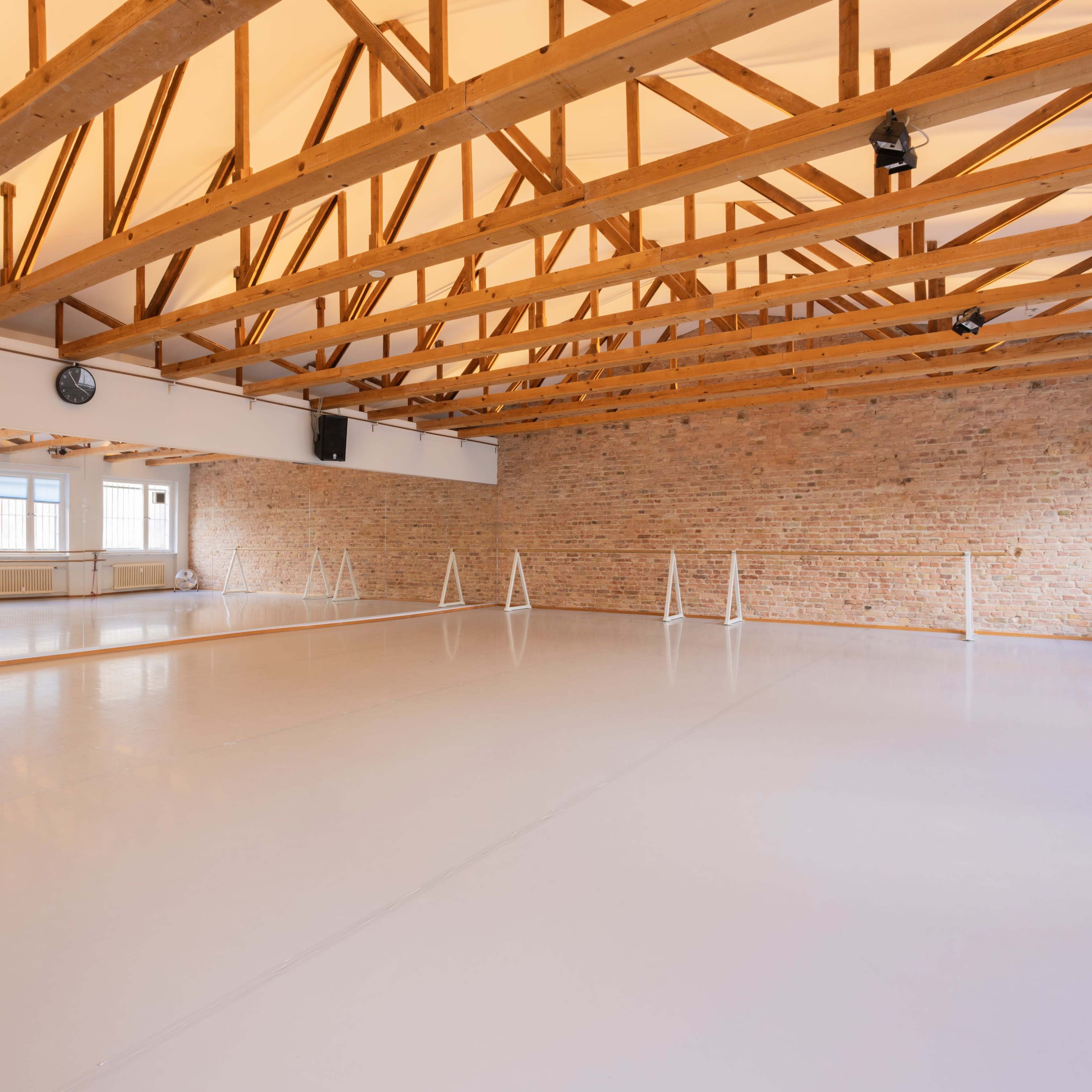 A spacious dance studio features polished white floors, exposed wooden beams, and large windows allowing natural light.