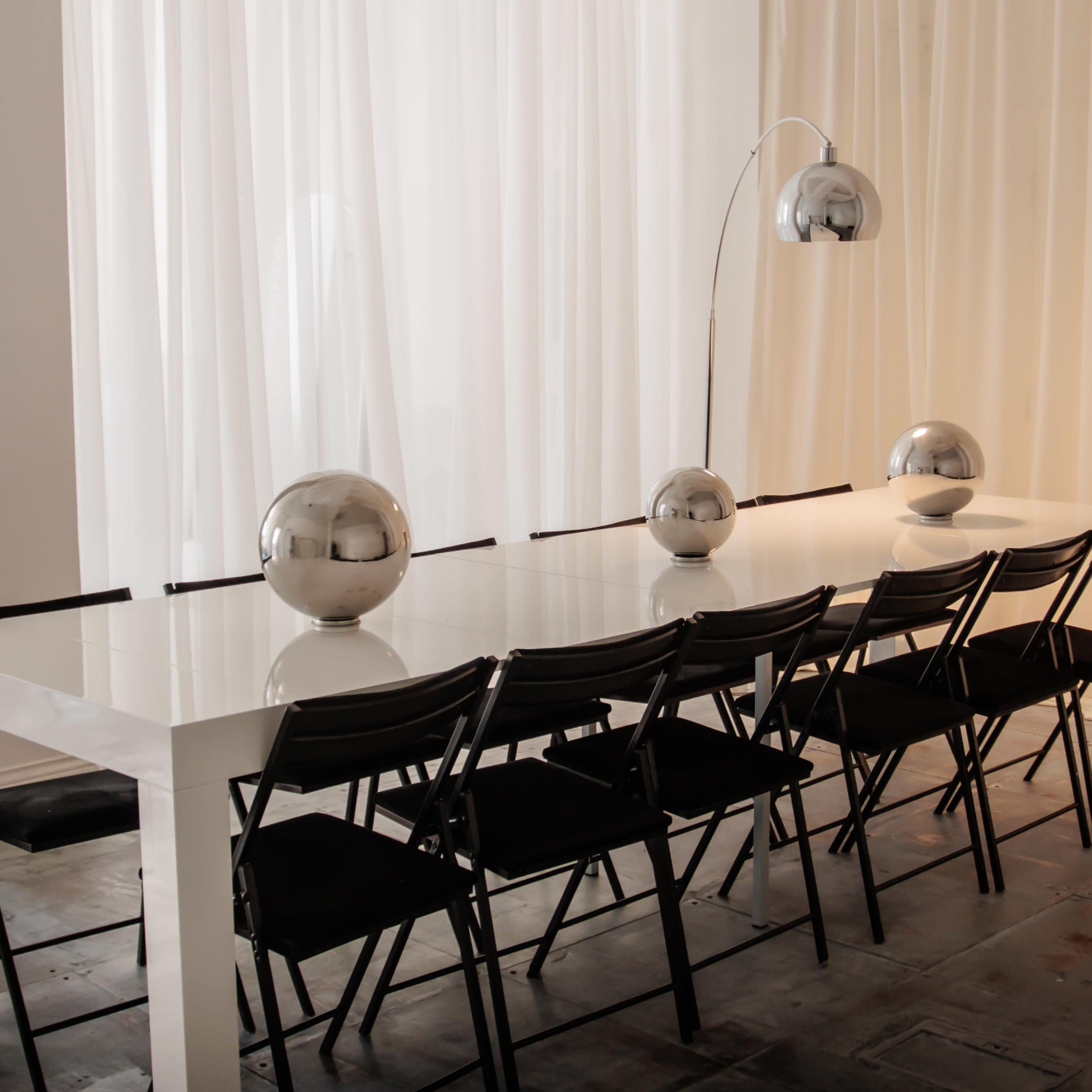 A long, white dining table is set with three metallic spheres, accompanied by black folding chairs and illuminated by an arc floor lamp.