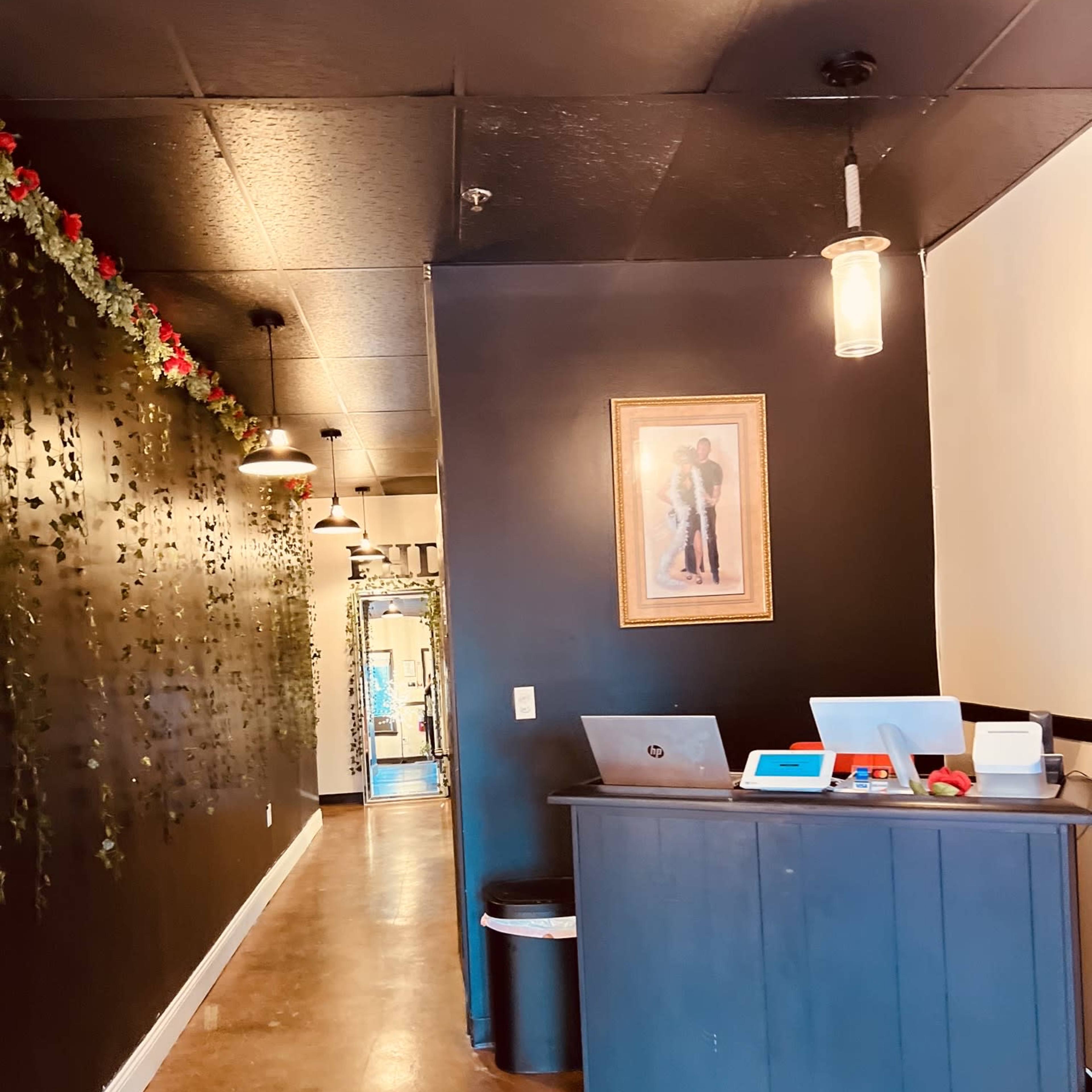 A narrow hallway with a reception desk on the left, decorated with a framed picture and illuminated by pendant lights, leading toward an entrance at the end.