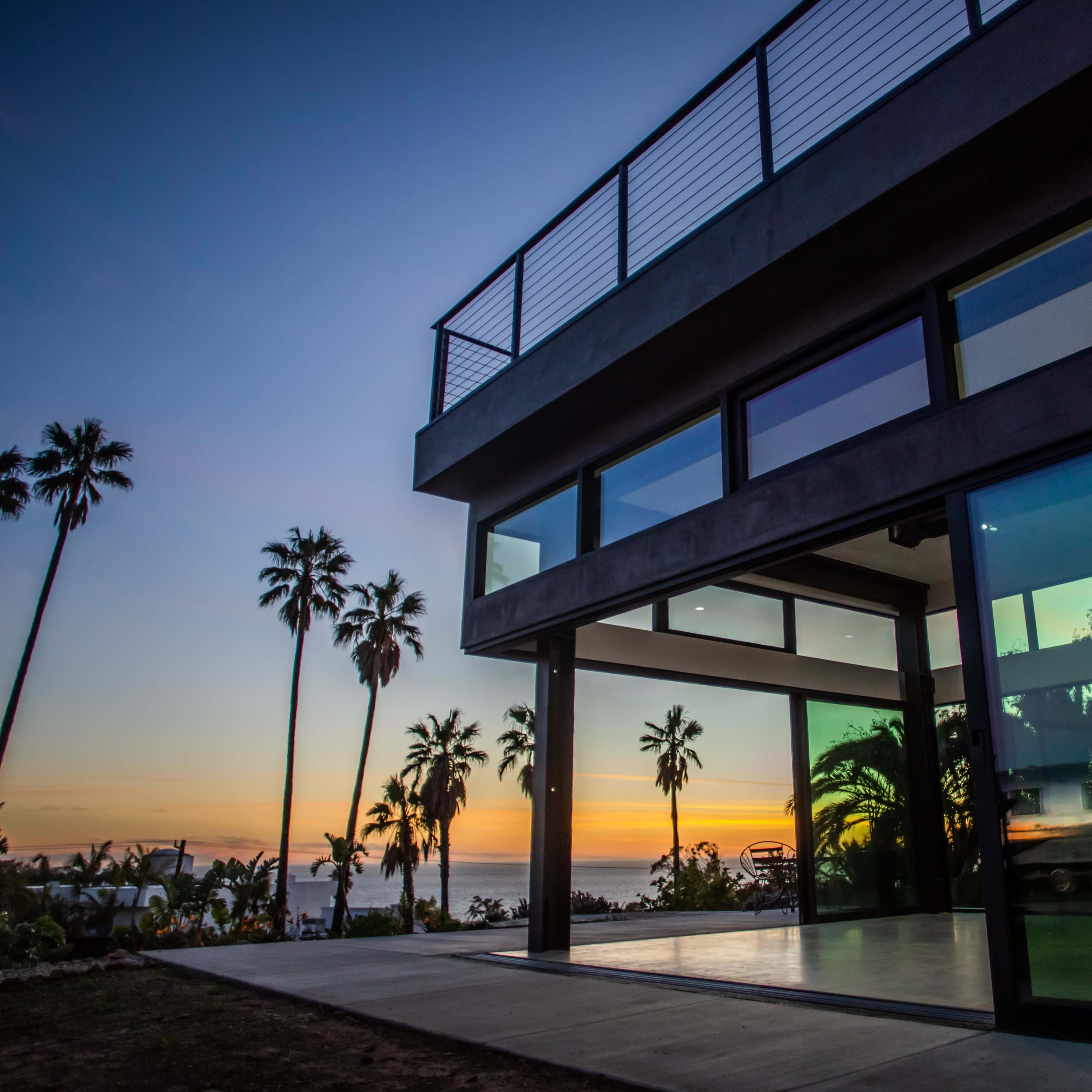 A modern house with large glass windows overlooks a sunset and palm trees.