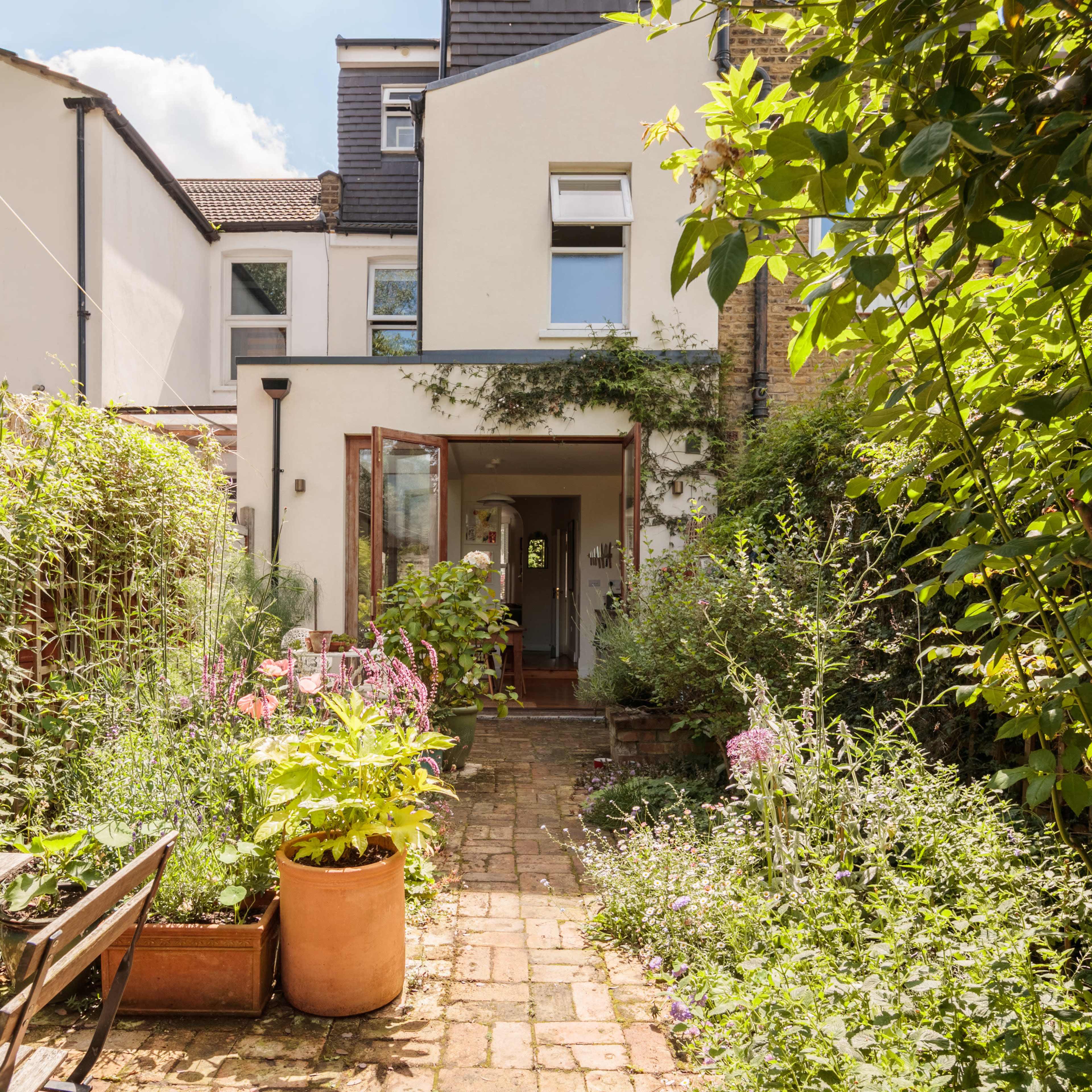 A brick pathway leads to an open doorway surrounded by lush greenery and flowering plants in a residential garden.