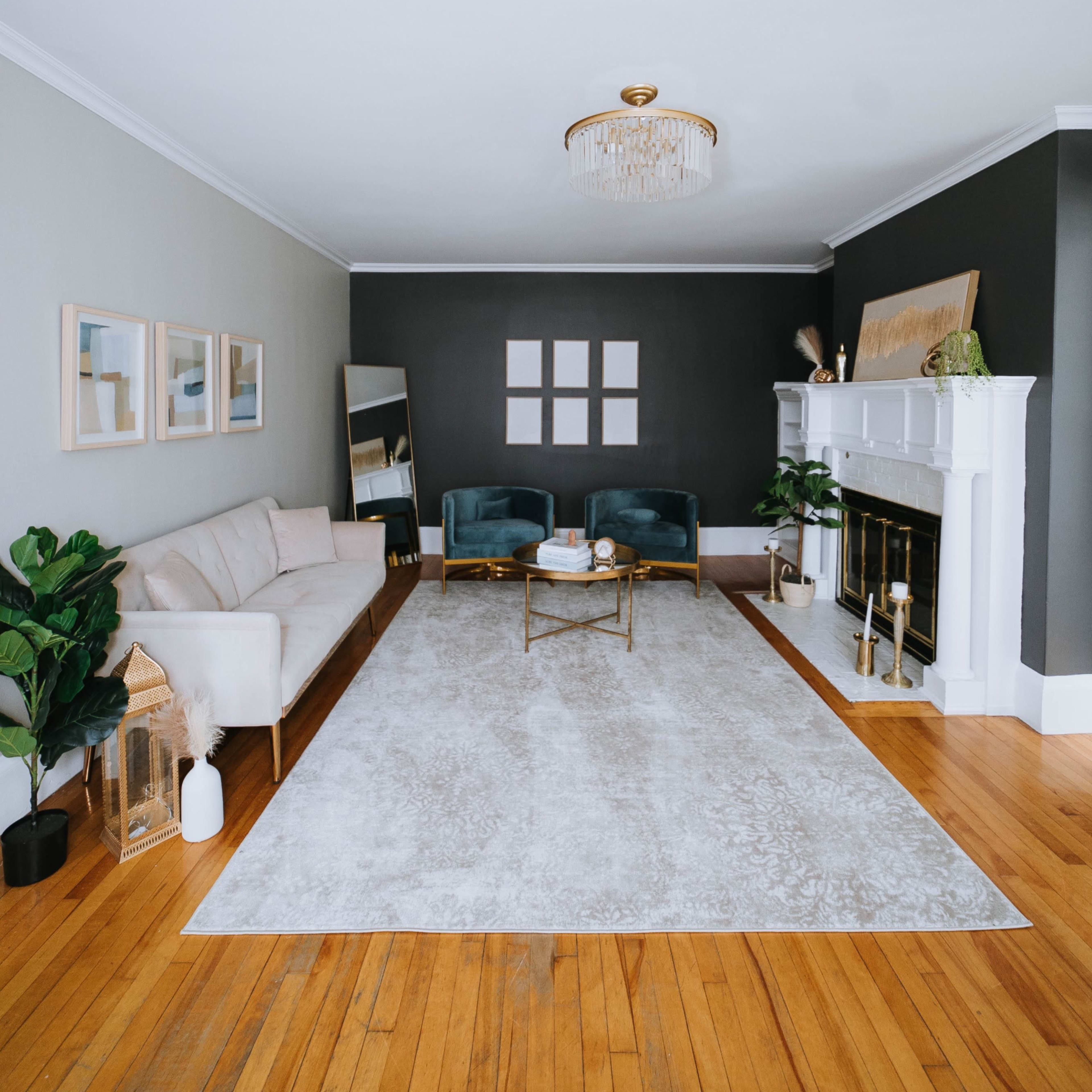 The room features a light-colored sofa, two green chairs, a coffee table on a large rug, and a fireplace against a dark wall, with framed pictures above.