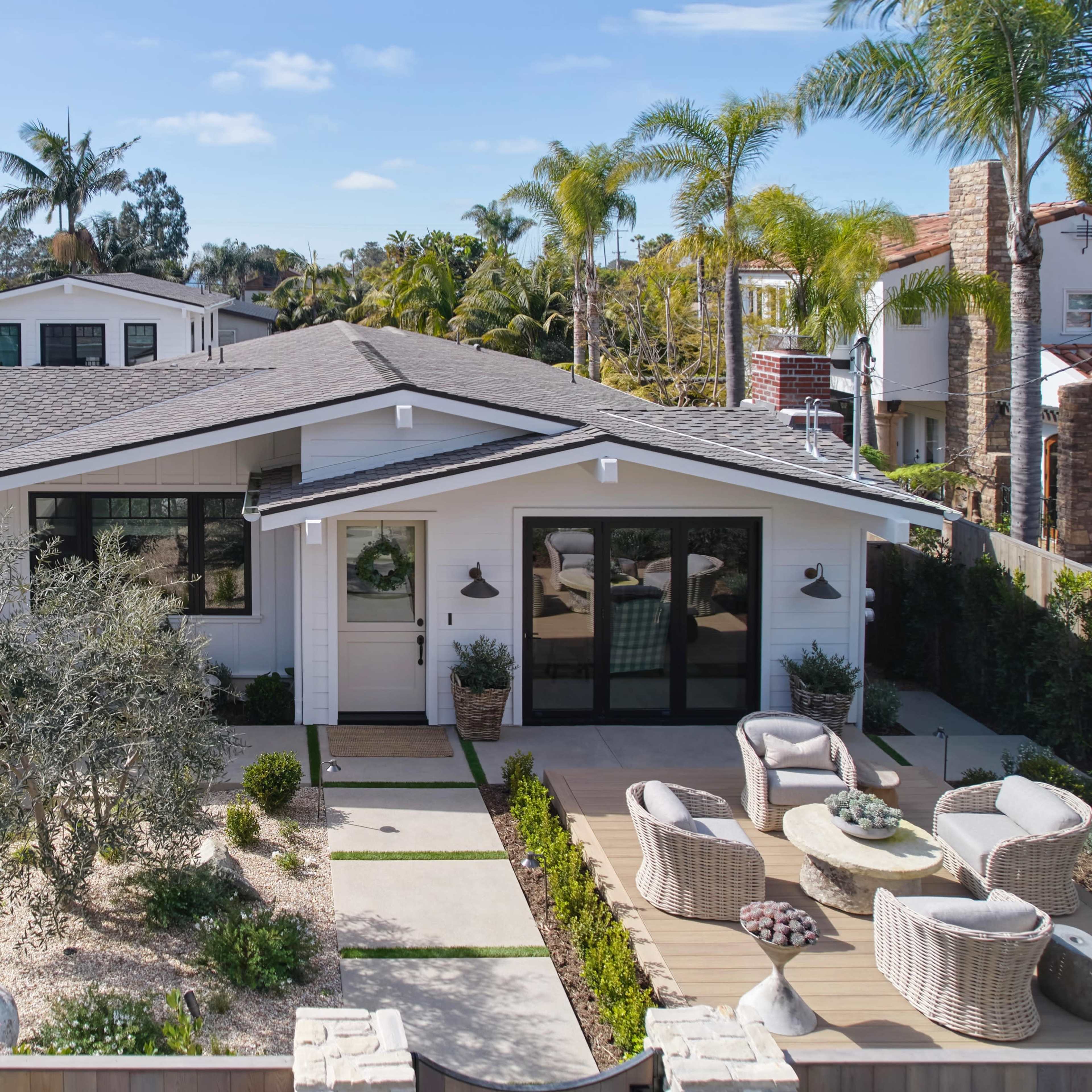 A modern single-story house with a gray roof, surrounded by palm trees and a landscaped yard featuring a patio area with outdoor furniture.