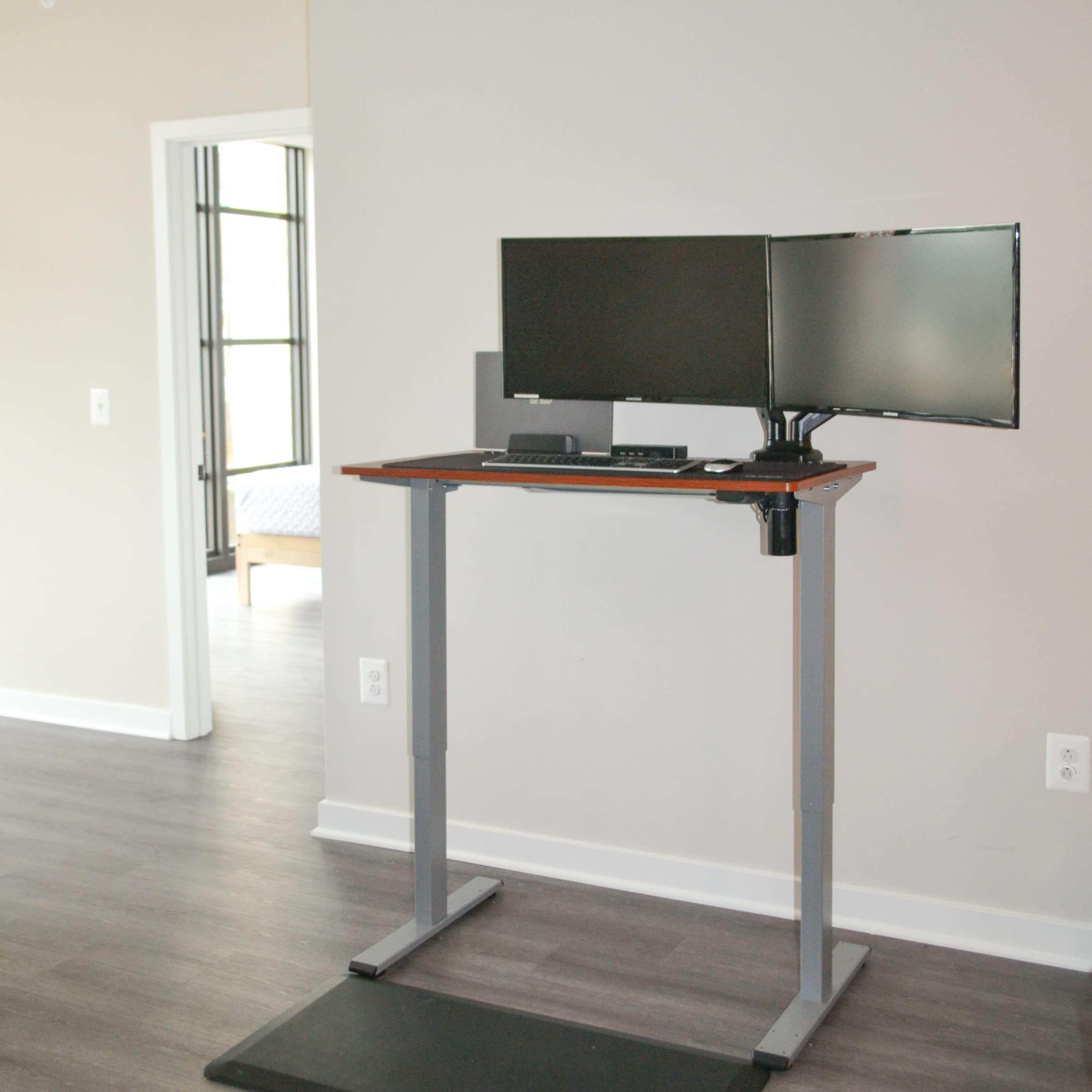 A standing desk with a dual-monitor setup is placed in a room with hardwood flooring and a doorway leading to another space.