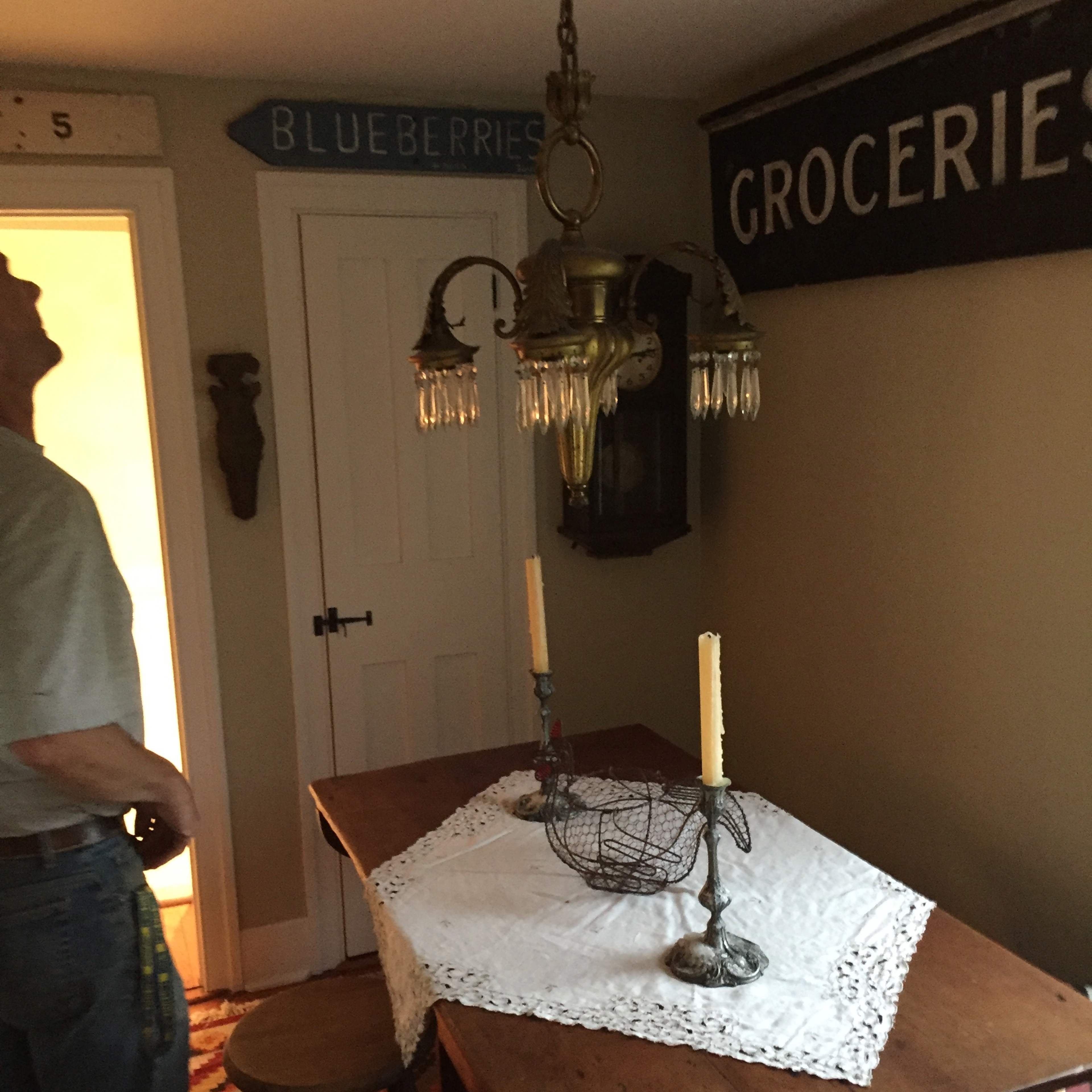 A man stands next to a wooden dining table set with a lace tablecloth and candles in a room featuring vintage decor, including a chandelier and a sign that reads "GROCERIES."