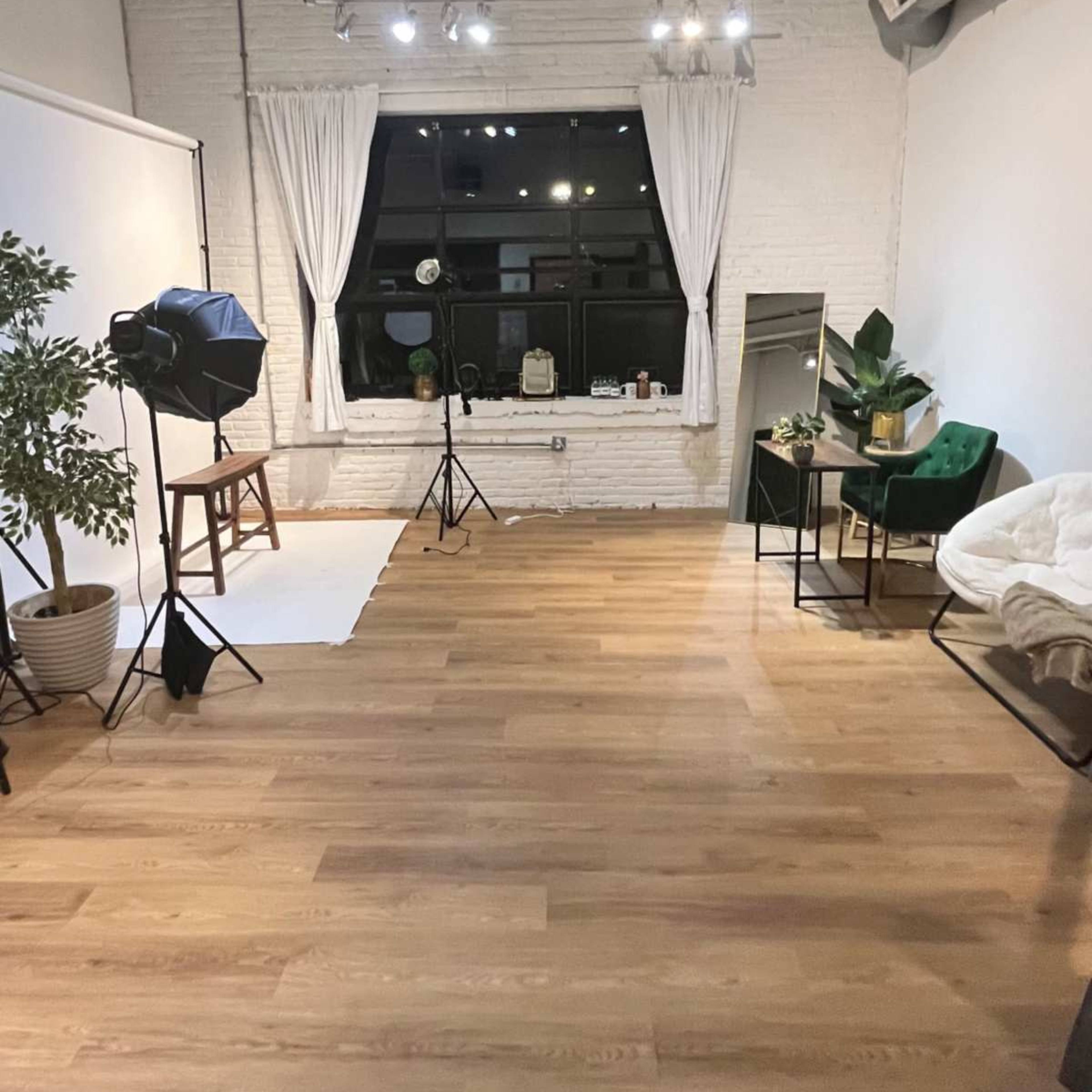 A spacious, well-lit studio with wooden flooring, a large window, photography equipment, and a cozy seating area adorned with plants.