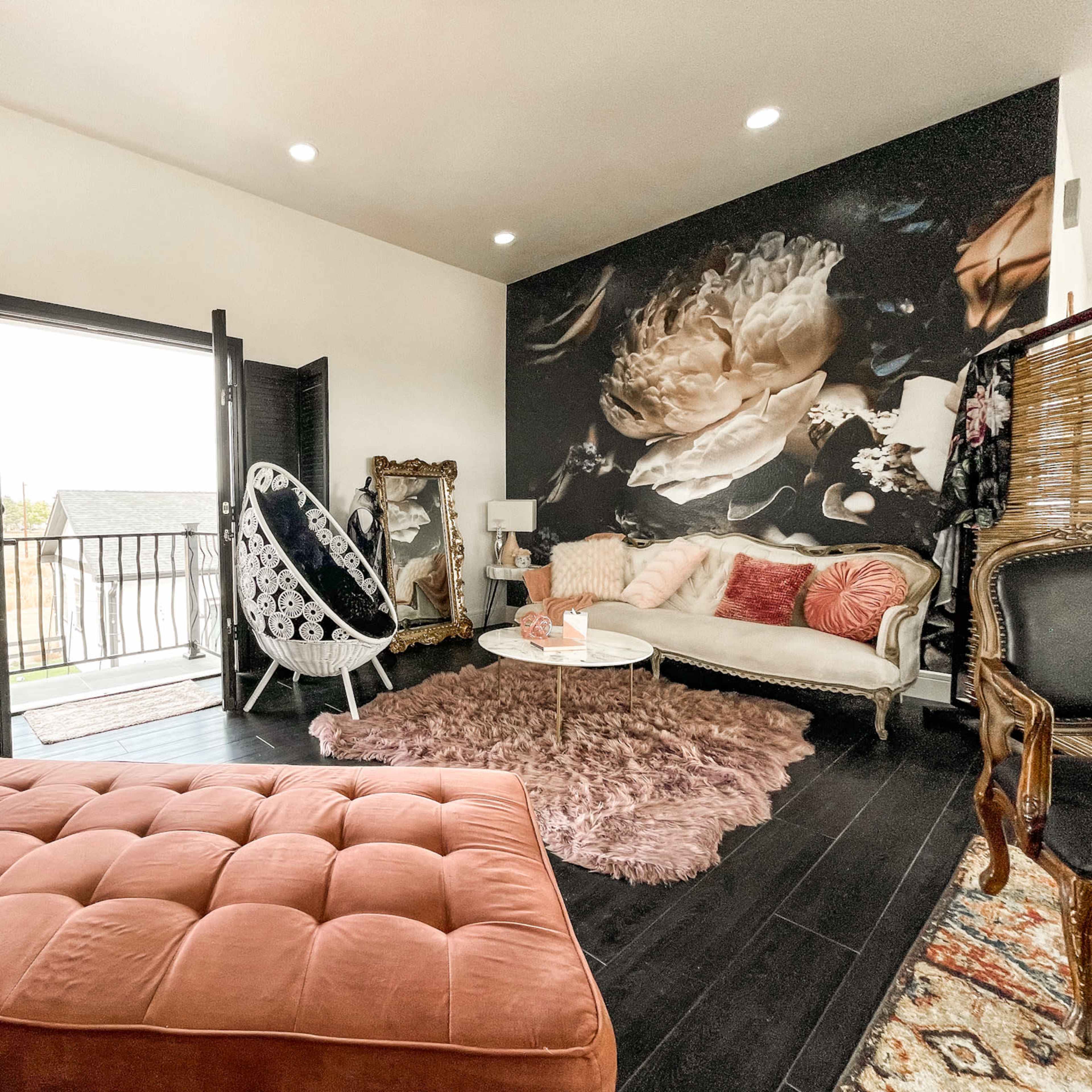 A stylish living room with a pink velvet sofa, a white hanging chair, and large windows opening to a balcony, accented by a floral mural on one wall.