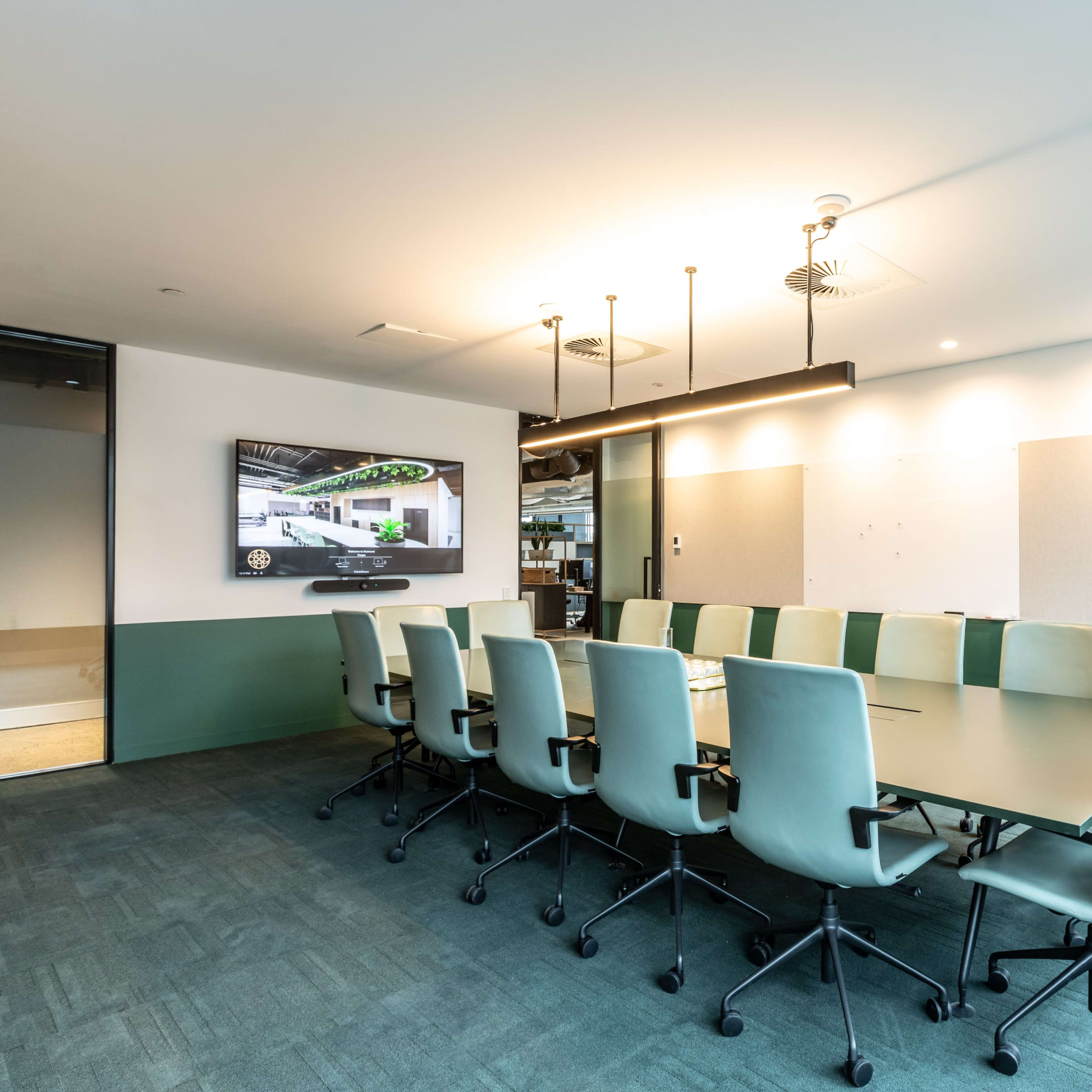 A conference room features a long table surrounded by green chairs and a large wall-mounted screen.
