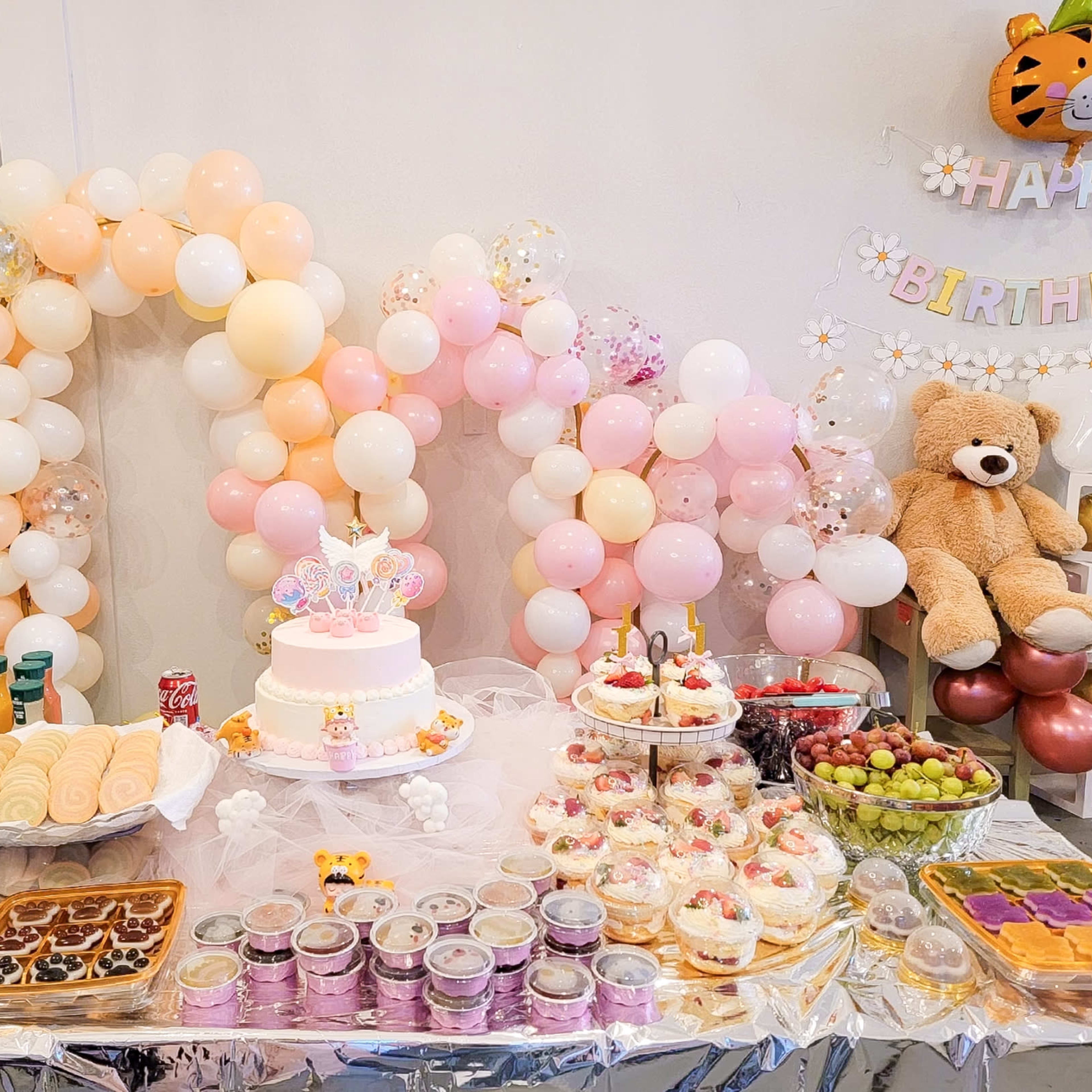 A dessert table is decorated with a birthday cake, pastries, snacks, and a colorful balloon arch.