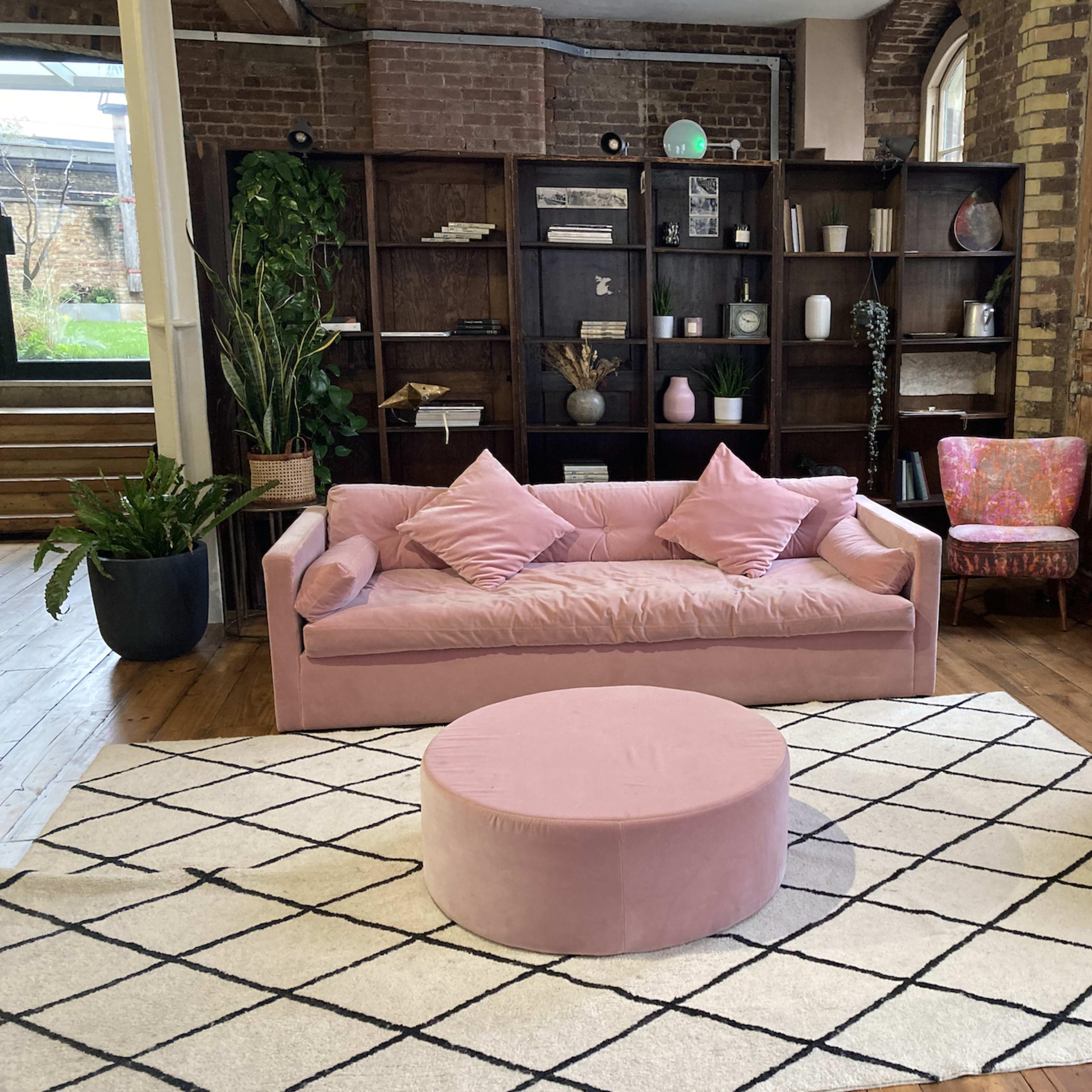 A modern living room featuring a pink couch, a round pink ottoman, a patterned rug, and a wooden bookshelf filled with various decorative items.