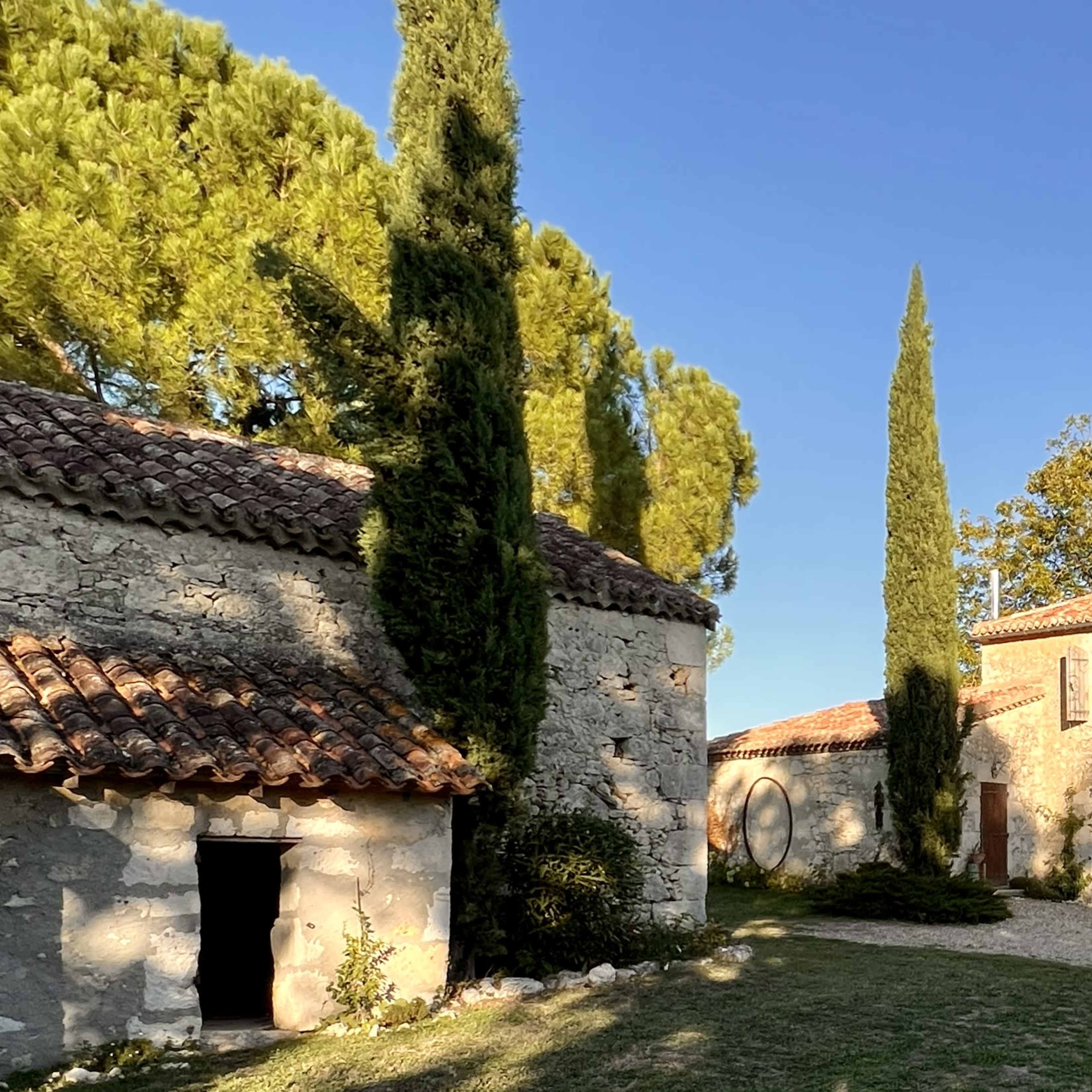 A stone house with a tiled roof surrounded by tall cypress trees and a gravel pathway.