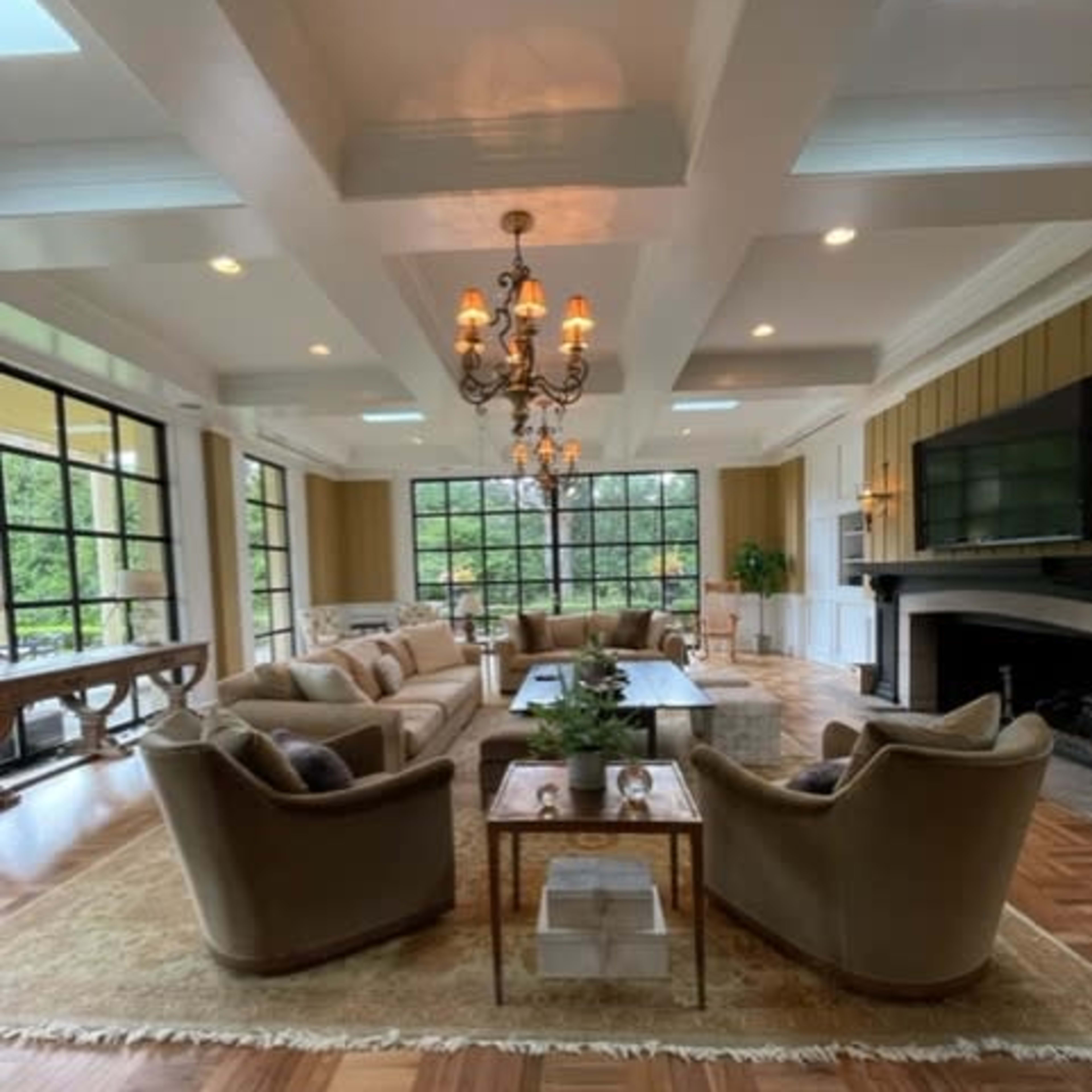 A spacious, well-lit living room with a large window, comfortable seating, and a modern chandelier.