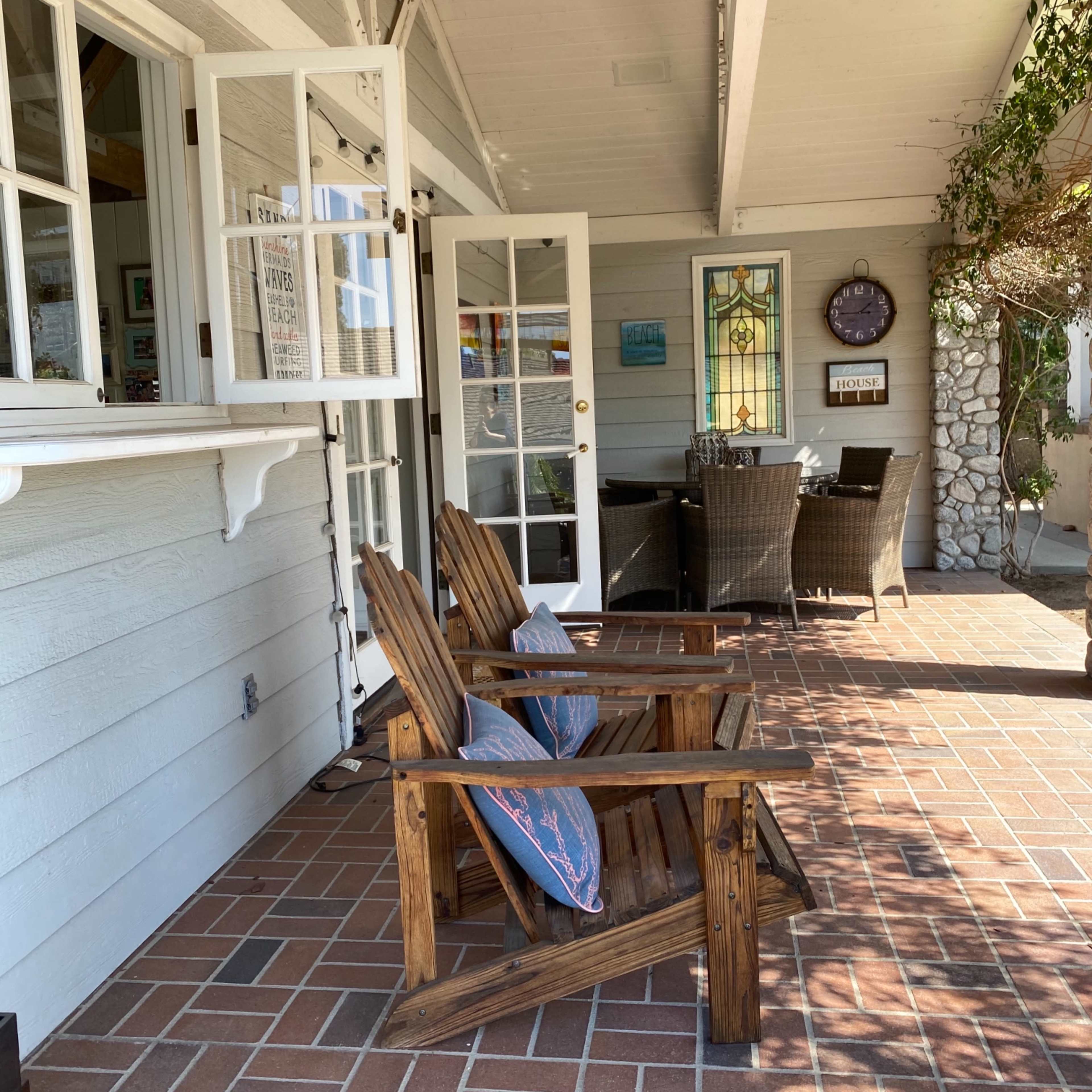 A cozy porch area with two wooden adirondack chairs facing a door, surrounded by a dining table and chairs under a shaded roof.