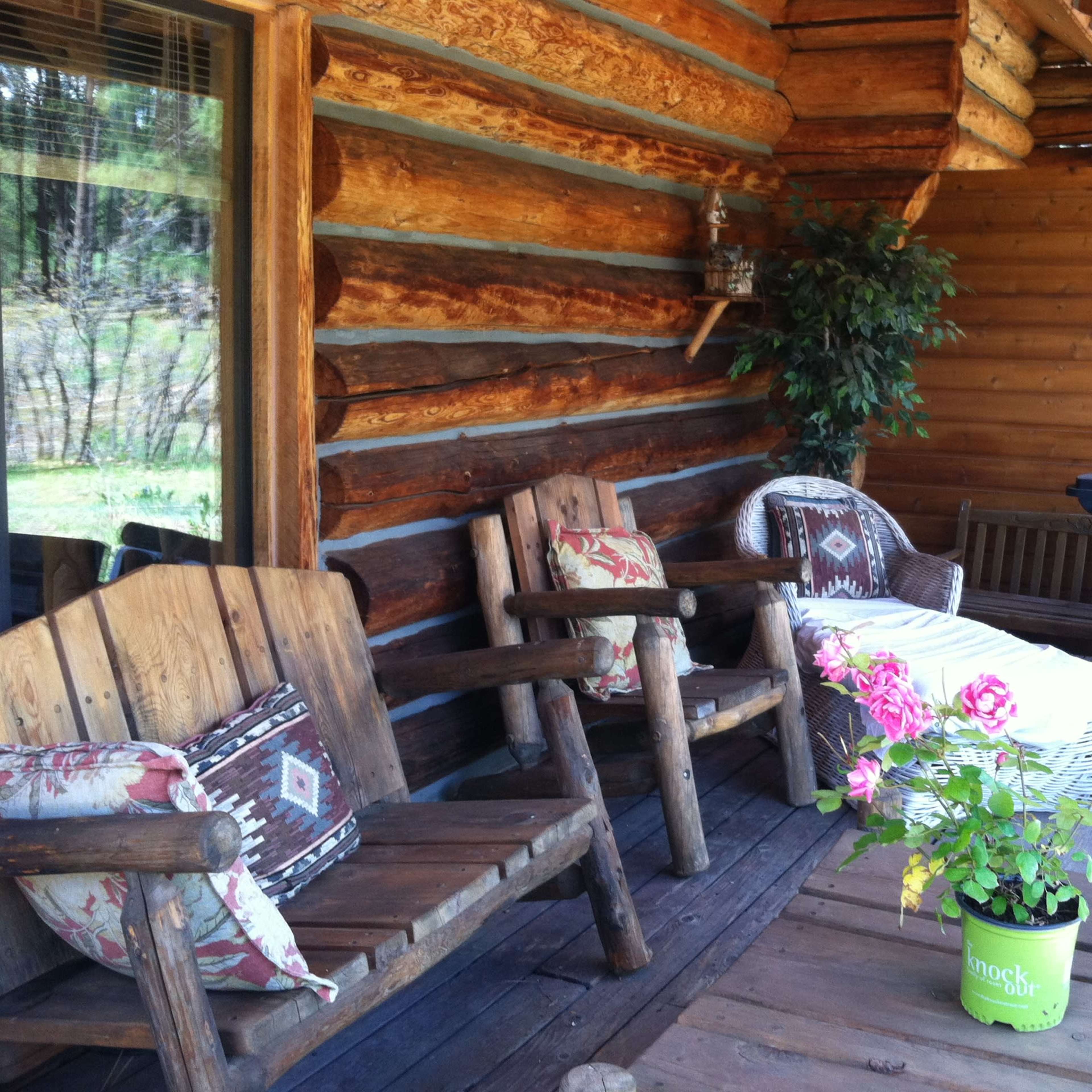 A wooden porch with two rustic chairs, a small table with a flower pot, and a grill in the background.